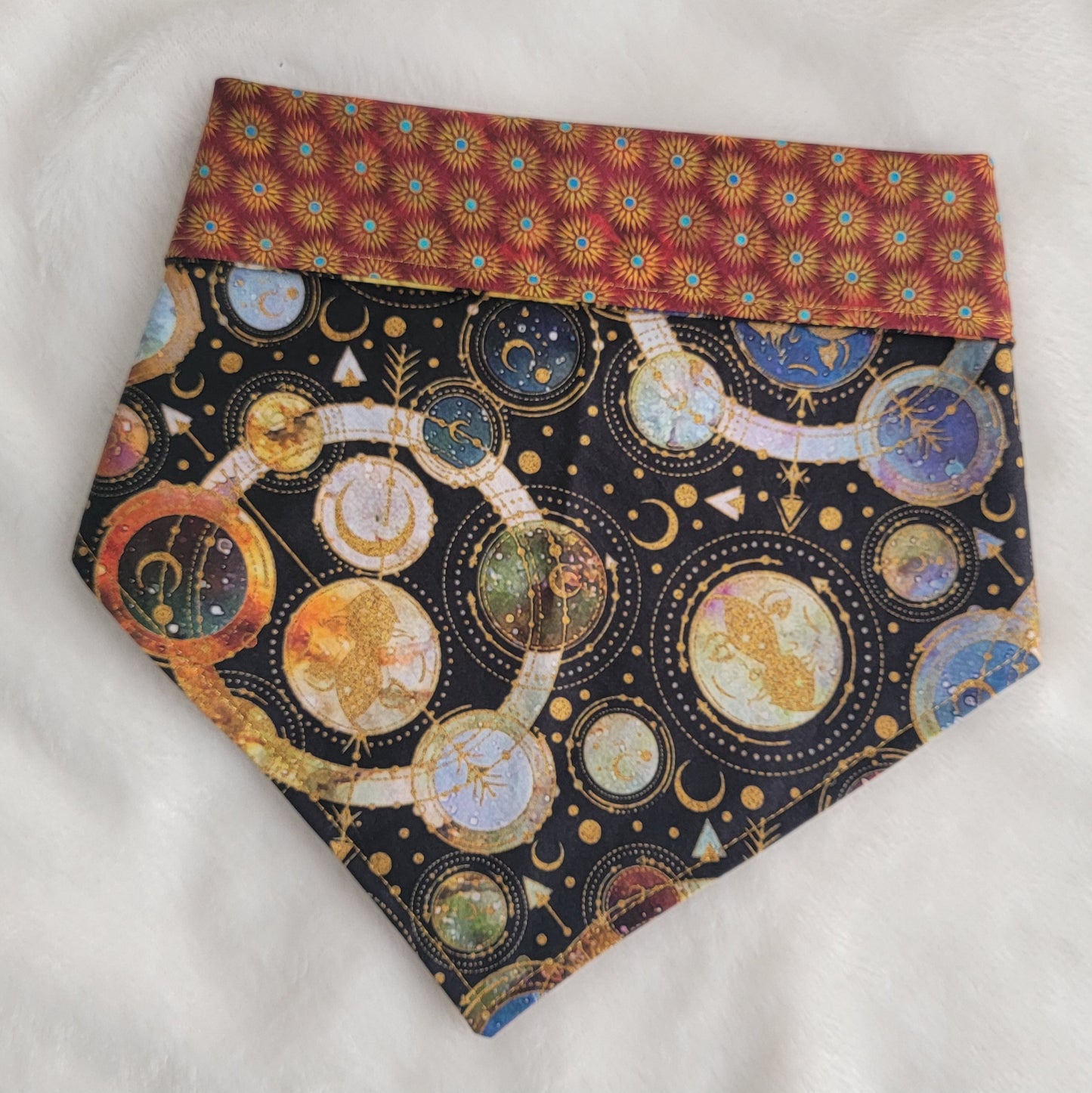 Reversible pet bandana, black and multicolor cosmic moons and planets geometric design with edge folded down to show red and gold sunburst print on reverse side.