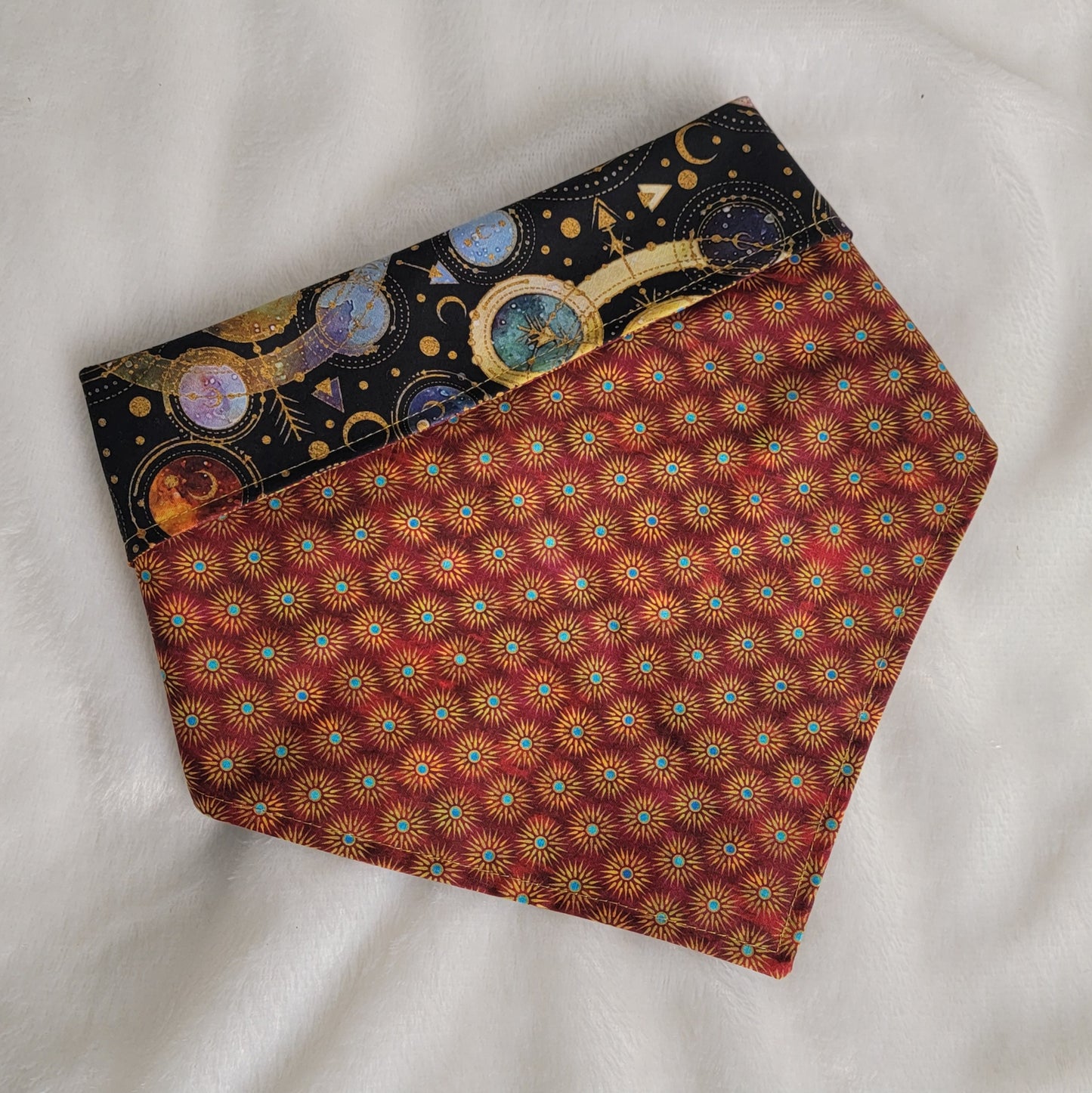 Reversible pet bandana, red and gold sunburst print with edge folded down to show black and multicolor cosmic moons and planets geometric design on reverse side.