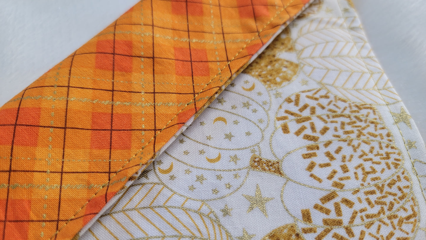 Close up of reversible pet bandana, folded showing one side white with gold pumpkins print and the other side bright orange plaid.
