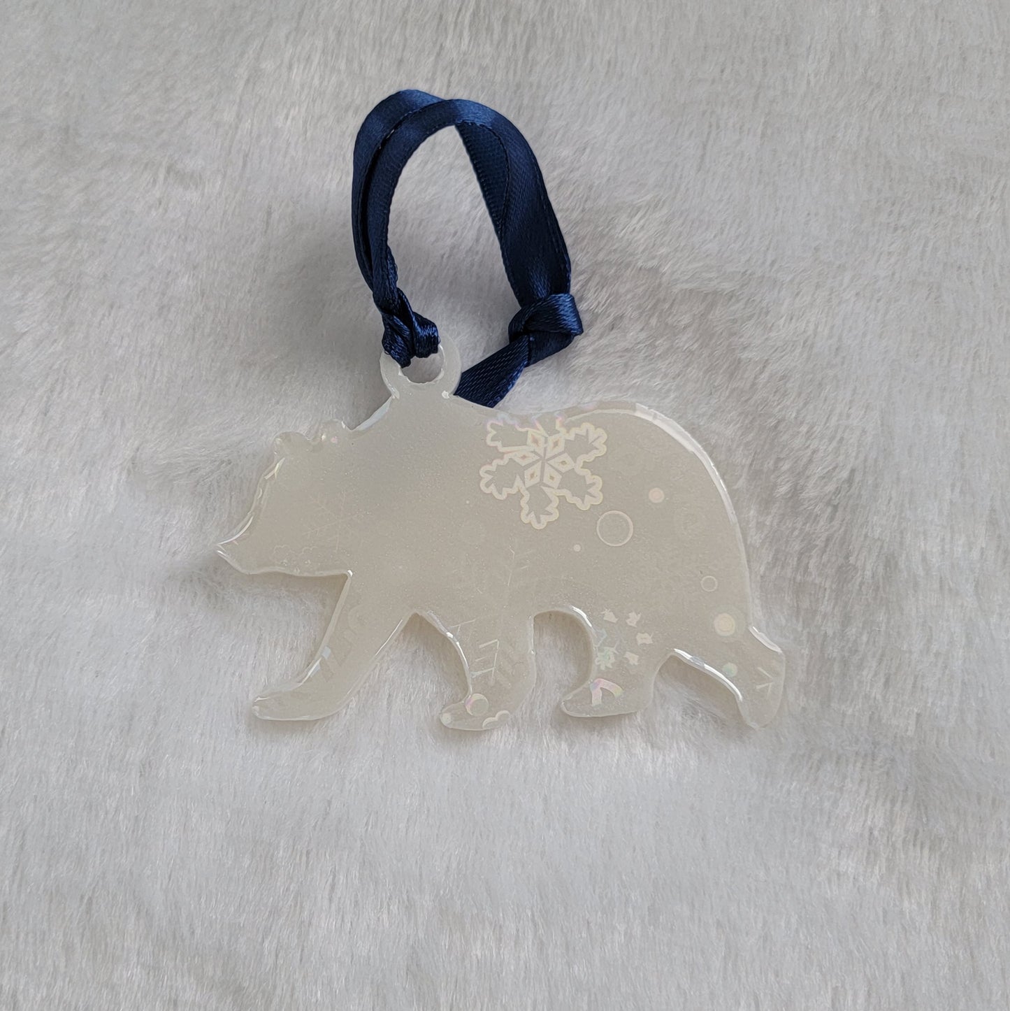 Bear shaped epoxy resin ornament, made with white pigment with a holographic snowflake print vinyl covering the entire piece and a dark blue ribbon attached.
