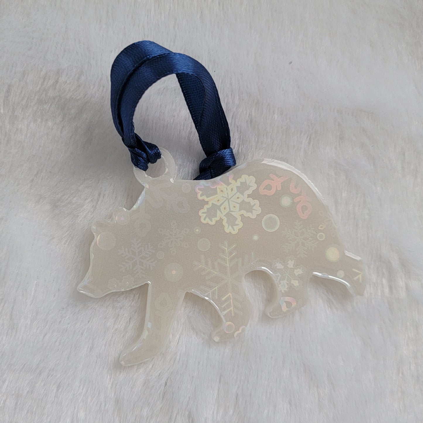 Bear shaped epoxy resin ornament, made with white pigment with a holographic snowflake print vinyl covering the entire piece and a dark blue ribbon attached.