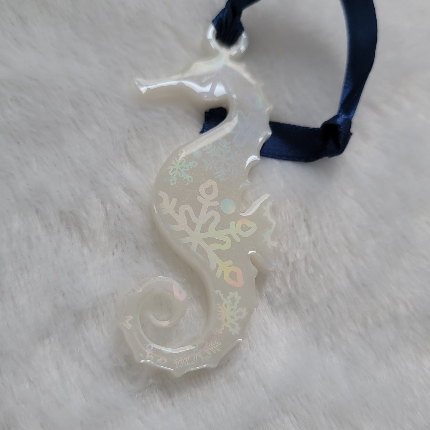 Seahorse shaped epoxy resin ornament, made with white pigment with a holographic snowflake print vinyl covering the entire piece and a dark blue ribbon attached.