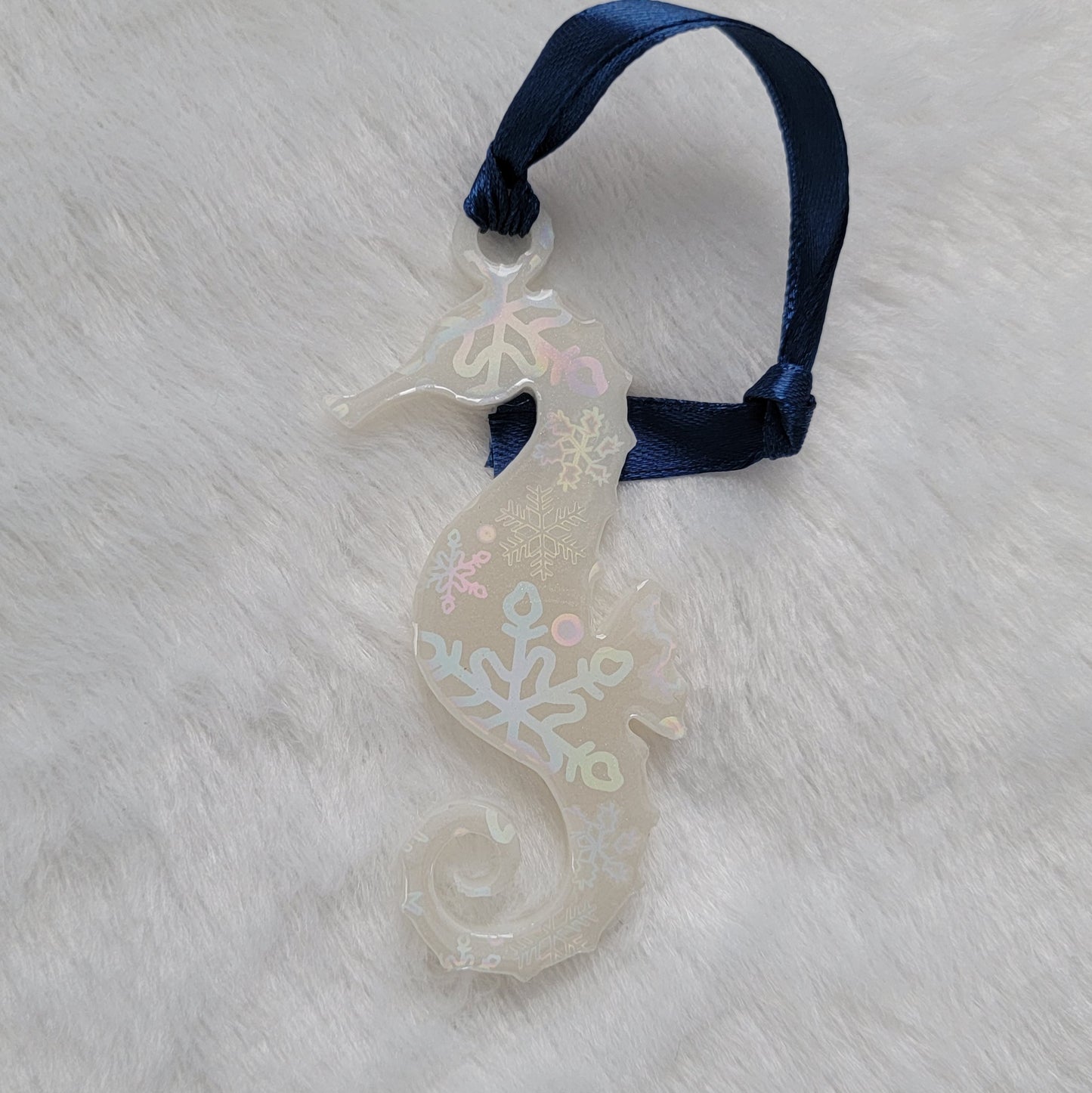 Seahorse shaped epoxy resin ornament, made with white pigment with a holographic snowflake print vinyl covering the entire piece and a dark blue ribbon attached.