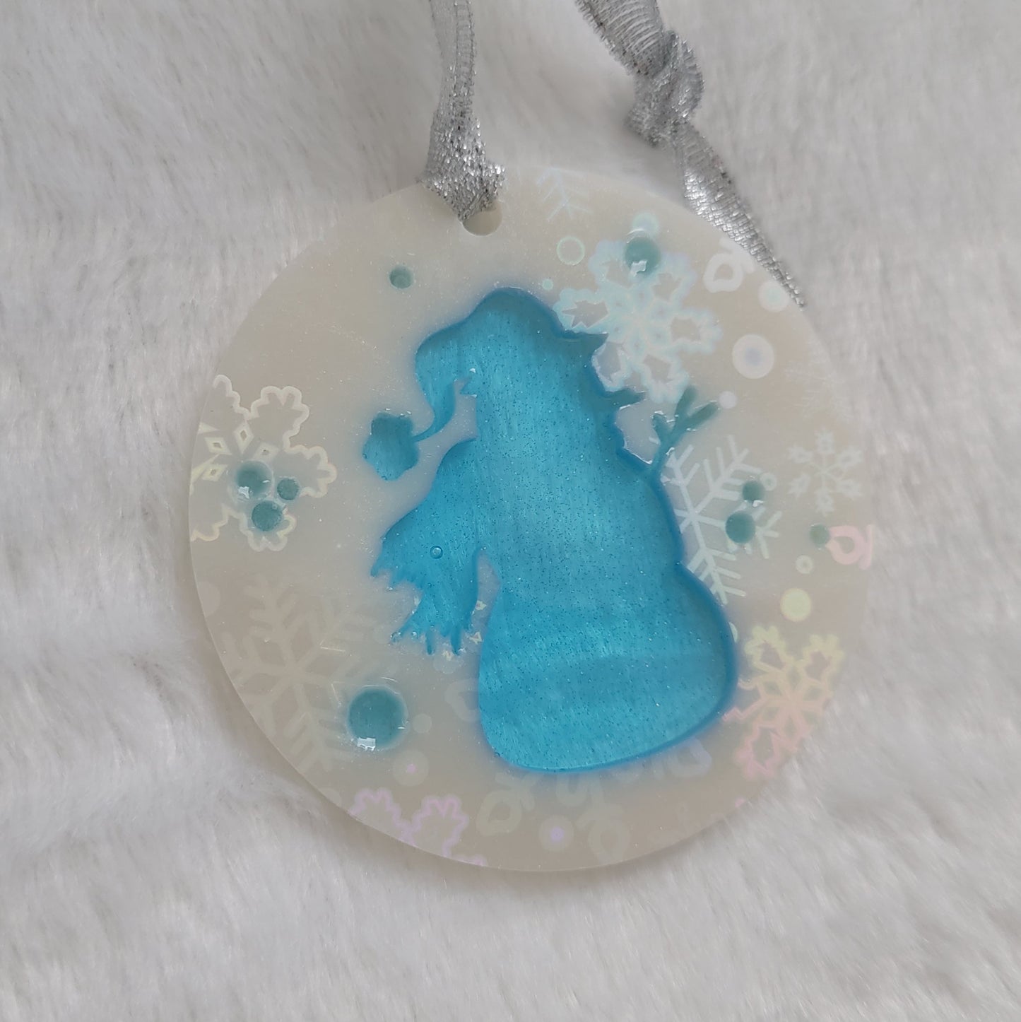 A round epoxy resin ornament that has a translucent blue snowman silhouette in the middle, made with white pigment with a holographic snowflake print vinyl covering the entire piece and a sparkling silver ribbon attached.
