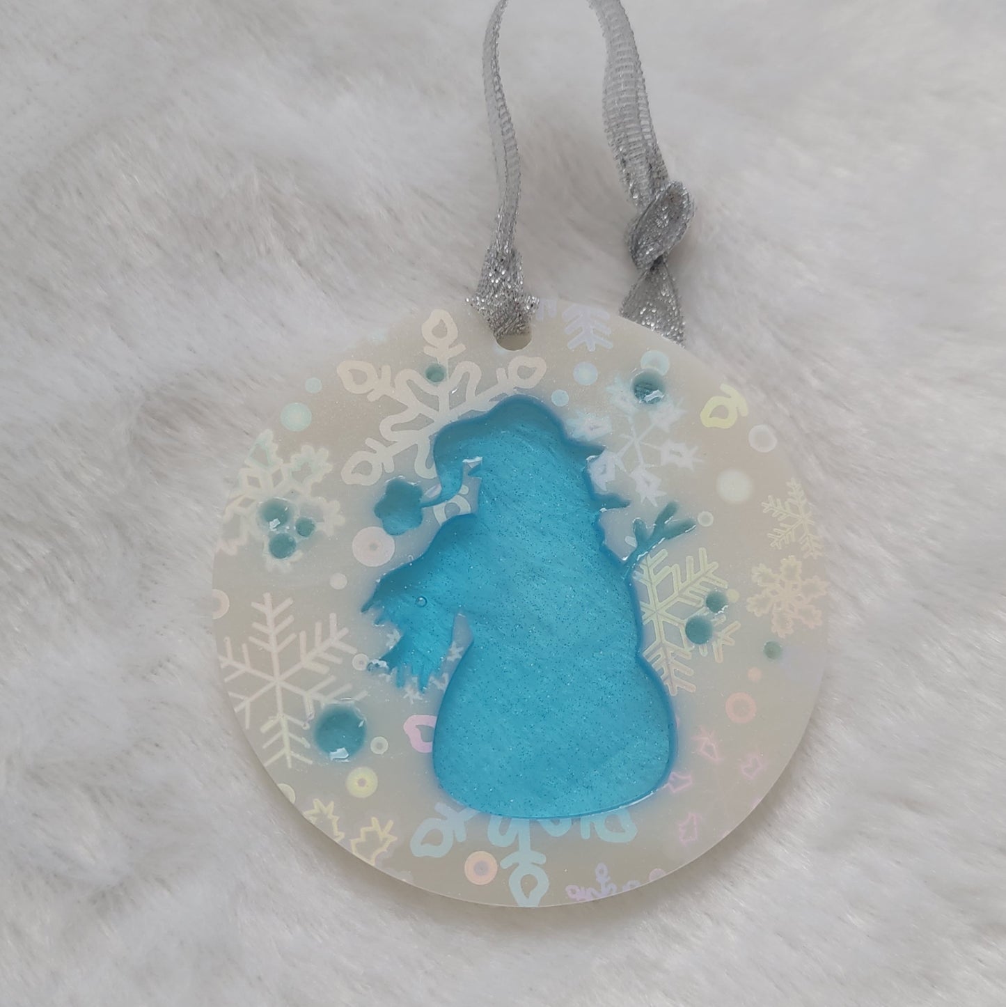 A round epoxy resin ornament that has a translucent blue snowman silhouette in the middle, made with white pigment with a holographic snowflake print vinyl covering the entire piece and a sparkling silver ribbon attached.