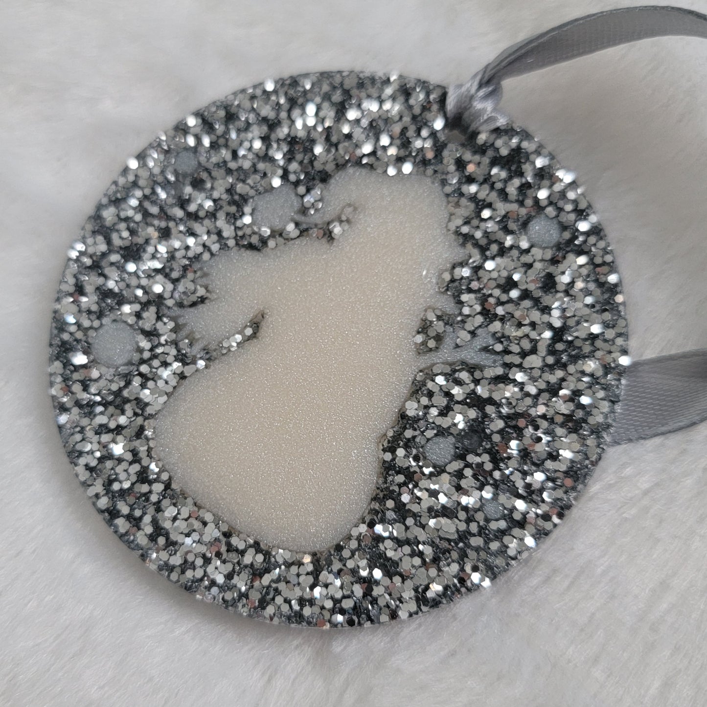 A round epoxy resin ornament that has a white snowman silhouette in the middle, made with bright silver glitter and a silver ribbon attached.