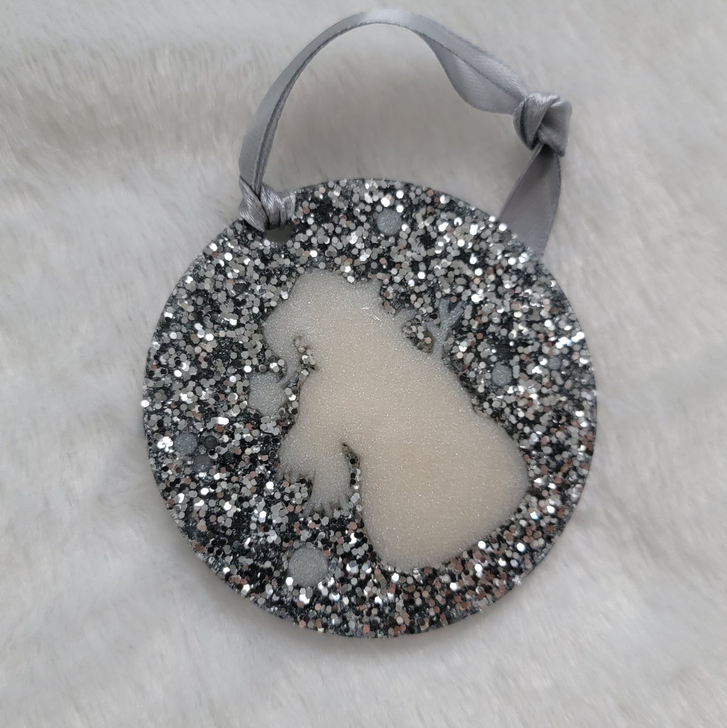 A round epoxy resin ornament that has a white snowman silhouette in the middle, made with bright silver glitter and a silver ribbon attached.