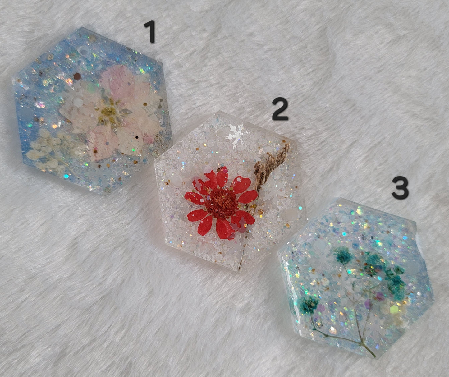 Three hexagon shaped glittery epoxy resin pieces that contain snowflake glitters and pressed flowers.