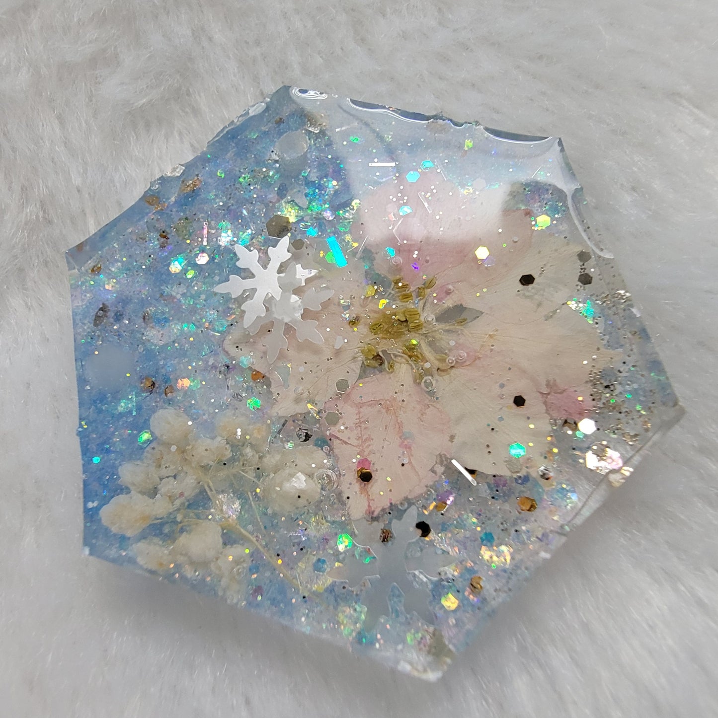 Glittery hexagon shaped epoxy resin piece containing snowflake glitters and a pink pressed flower and white pom blossoms.