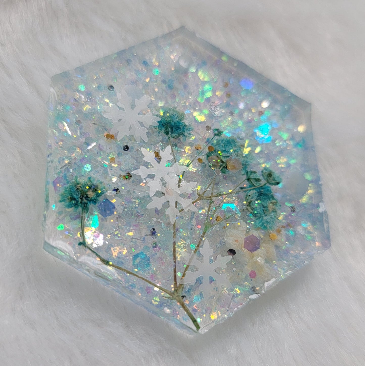 Glittery hexagon shaped epoxy resin piece containing snowflake glitters and blue flower stalks and white pom blossoms.