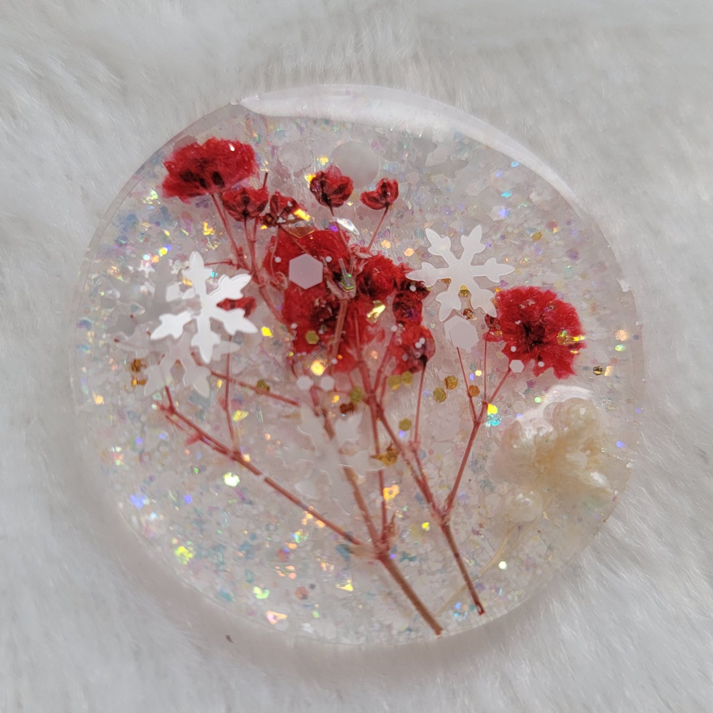 Glittery round epoxy resin piece containing snowflake glitters and red pressed flower stalks and white pom blossoms.