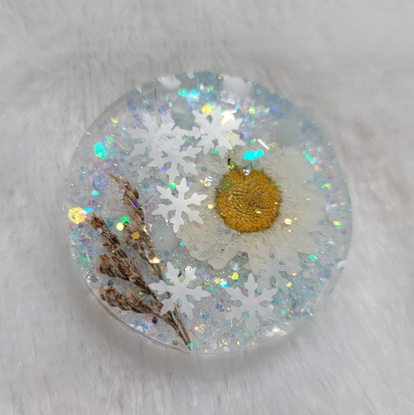 Glittery round epoxy resin piece containing snowflake glitters and a white and yellow daisy and brown grass blossom stalks.