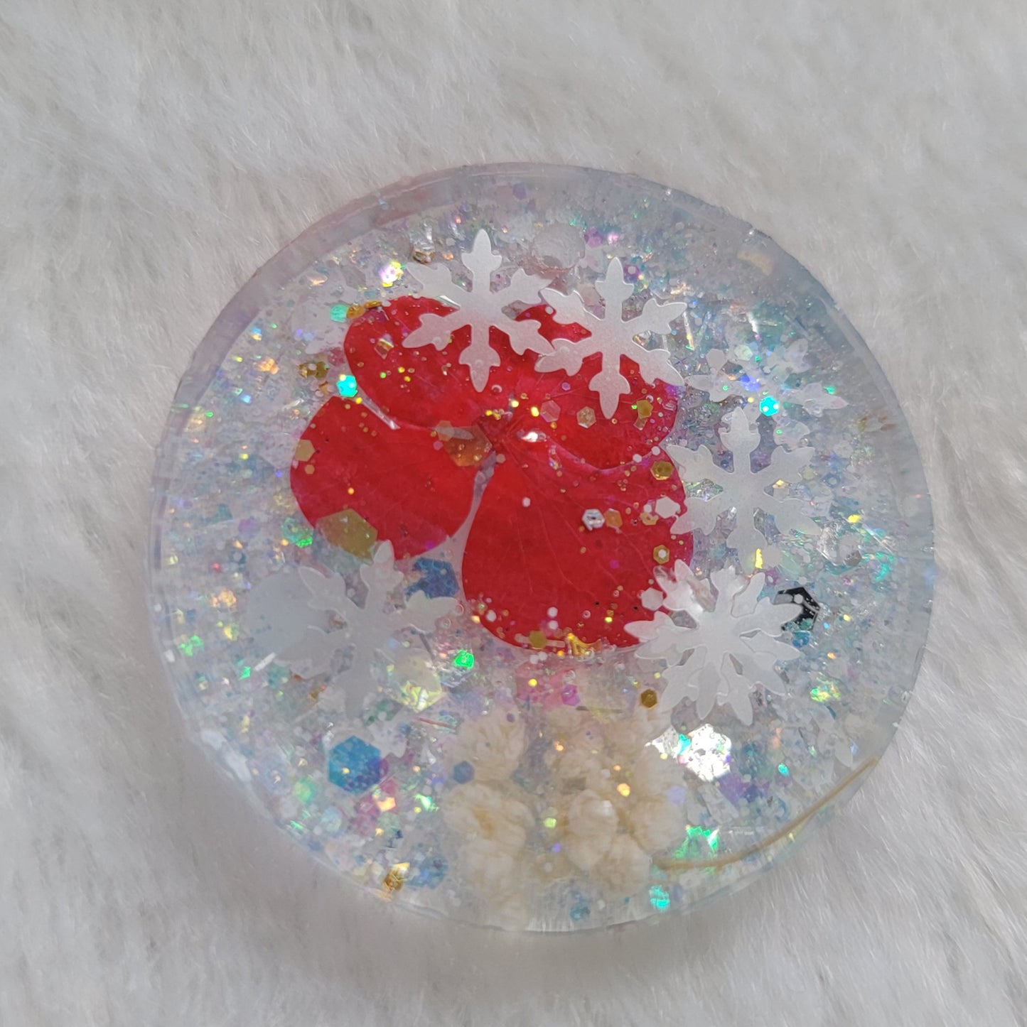 Glittery round epoxy resin piece containing snowflake glitters and bright red pressed flower and white pom blossoms.