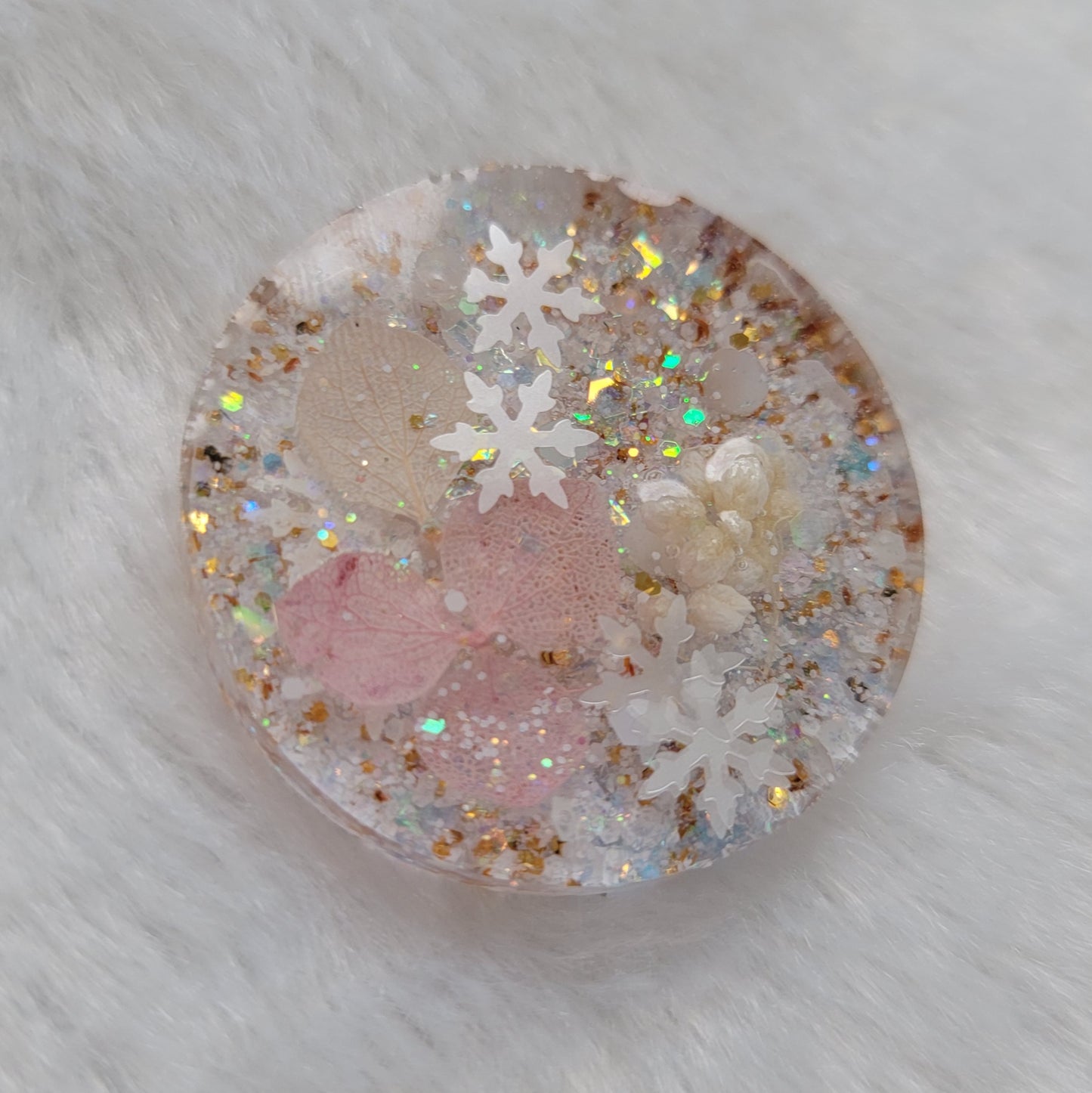 Glittery round epoxy resin piece containing snowflake glitters and pink pressed flowers and white pom blossoms.