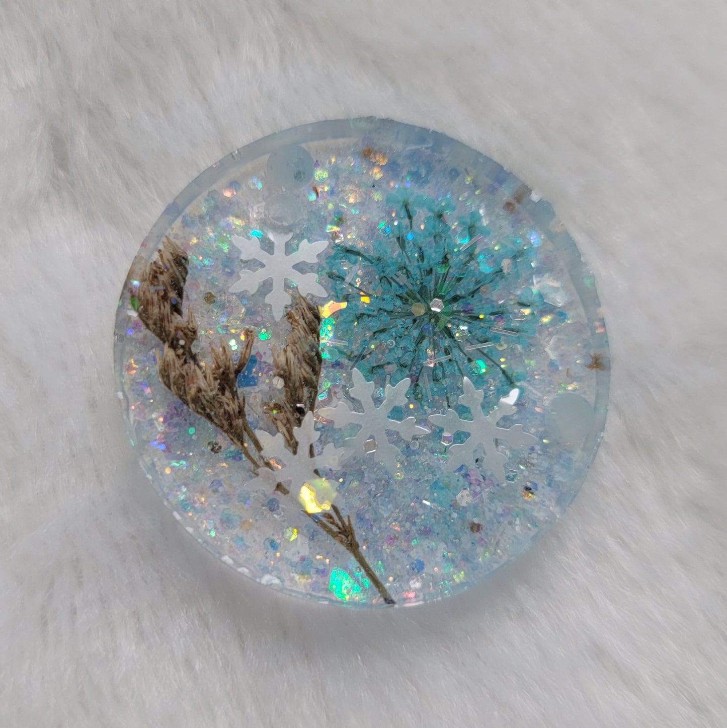 Glittery round epoxy resin piece containing snowflake glitters and a blue pressed flower and brown grass blossom stalks.