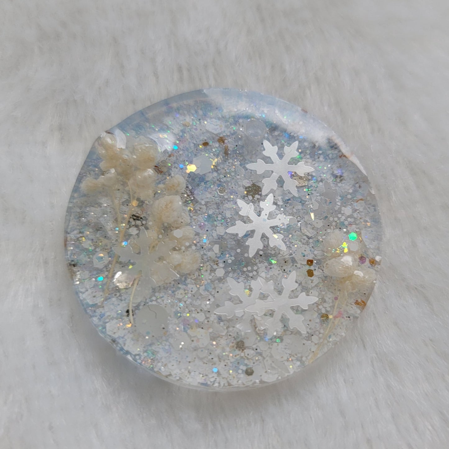 Glittery round epoxy resin piece containing snowflake glitters and white pom blossoms.