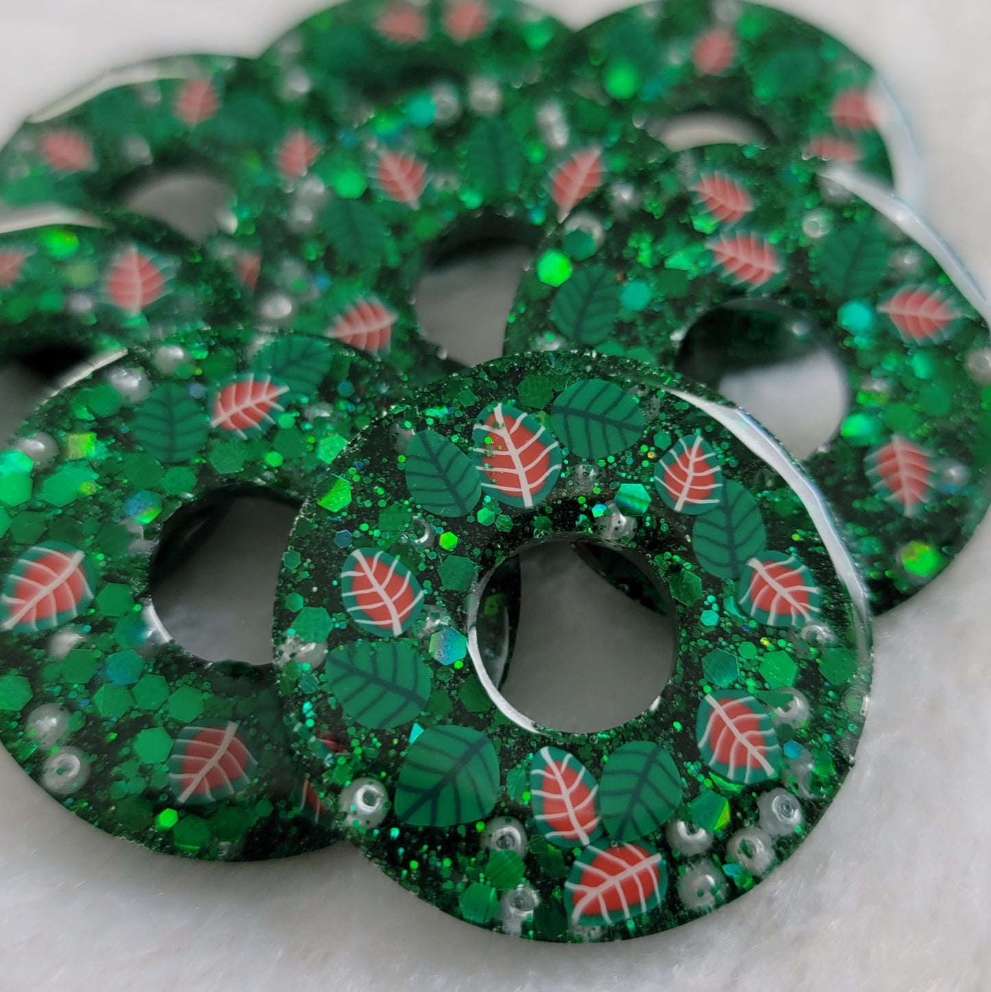 Eight small epoxy resin pieces made to look like holiday wreaths. They are wreath shaped, contain green glitter, small pearly white beads, and have green and red polymer leaves.