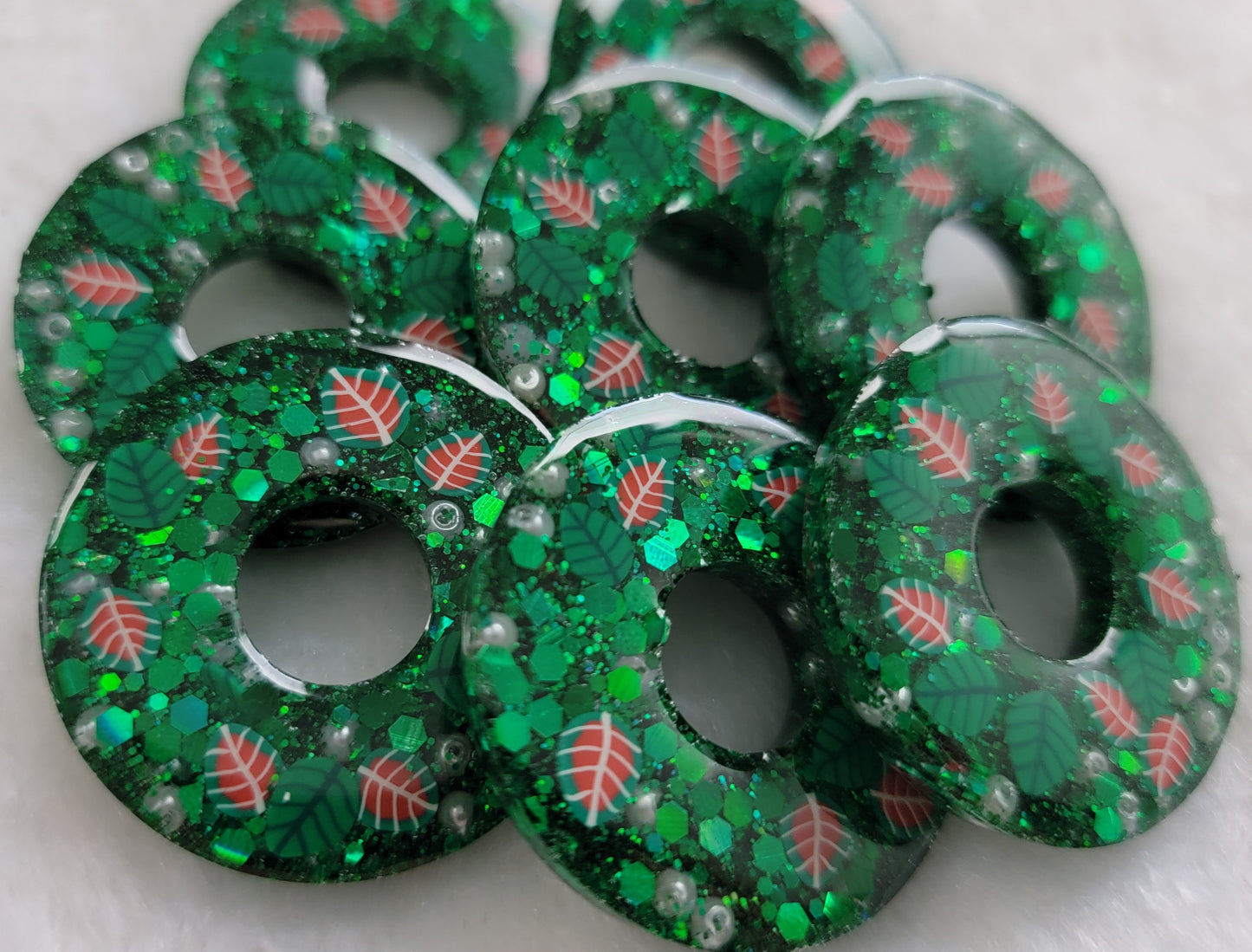 Eight small epoxy resin pieces made to look like holiday wreaths. They are wreath shaped, contain green glitter, small pearly white beads, and have green and red polymer leaves.