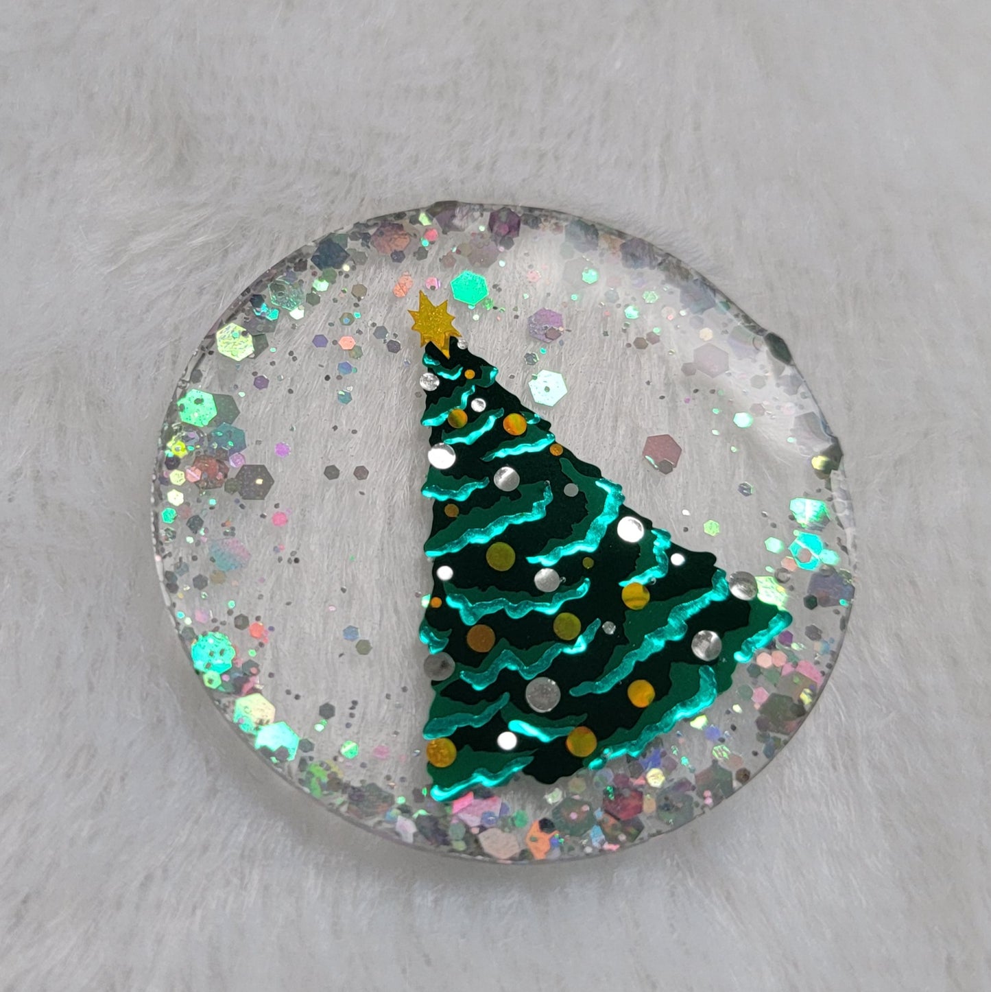 Round epoxy resin piece, base is transparent with silver holographic glitter around the edges, in the center there is a Christmas tree design made with vinyl in shades of green, silver and gold.