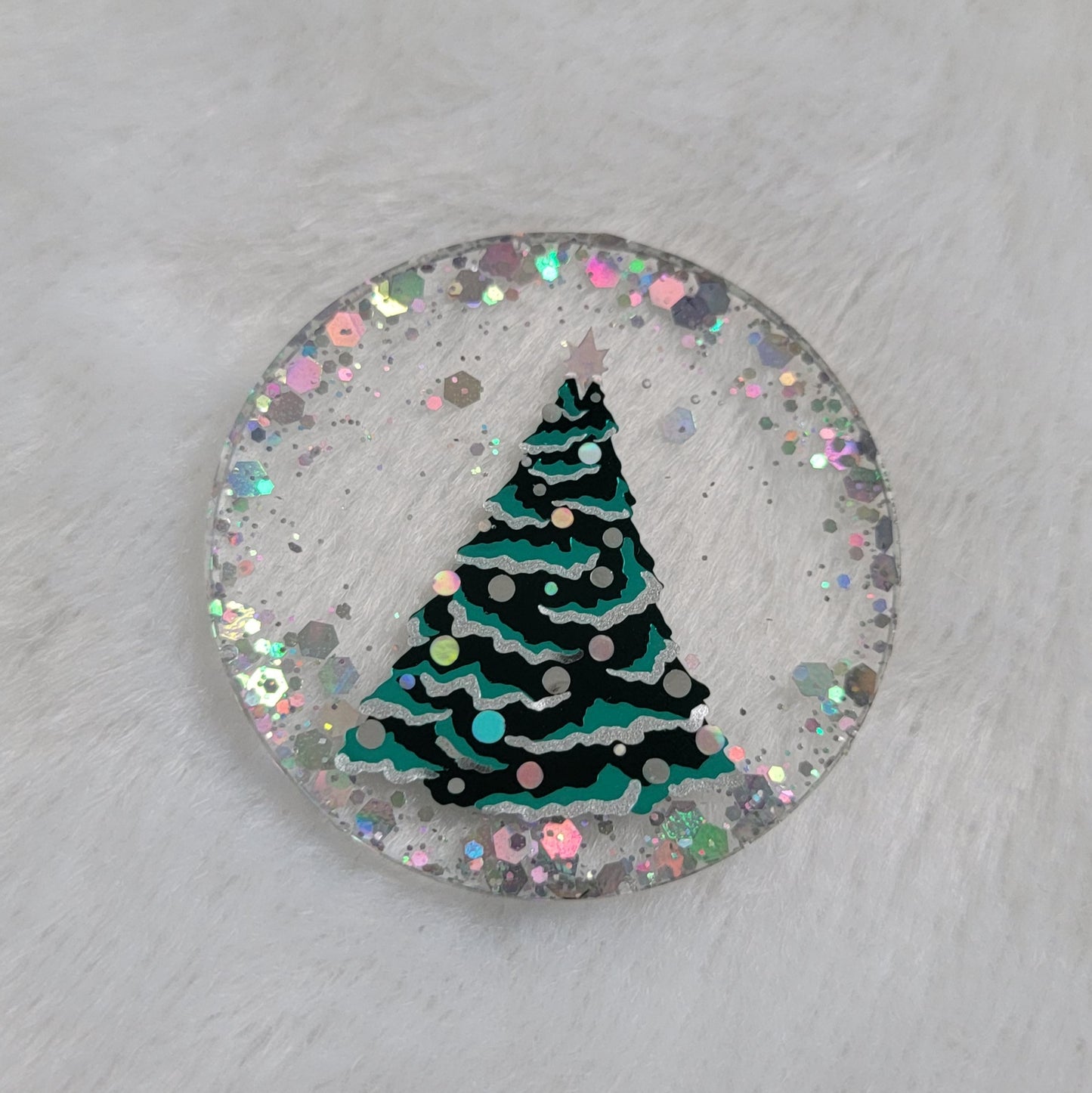 Round epoxy resin piece, base is transparent with silver holographic glitter around the edges, in the center there is a Christmas tree design made with vinyl in shades of green and silver.
