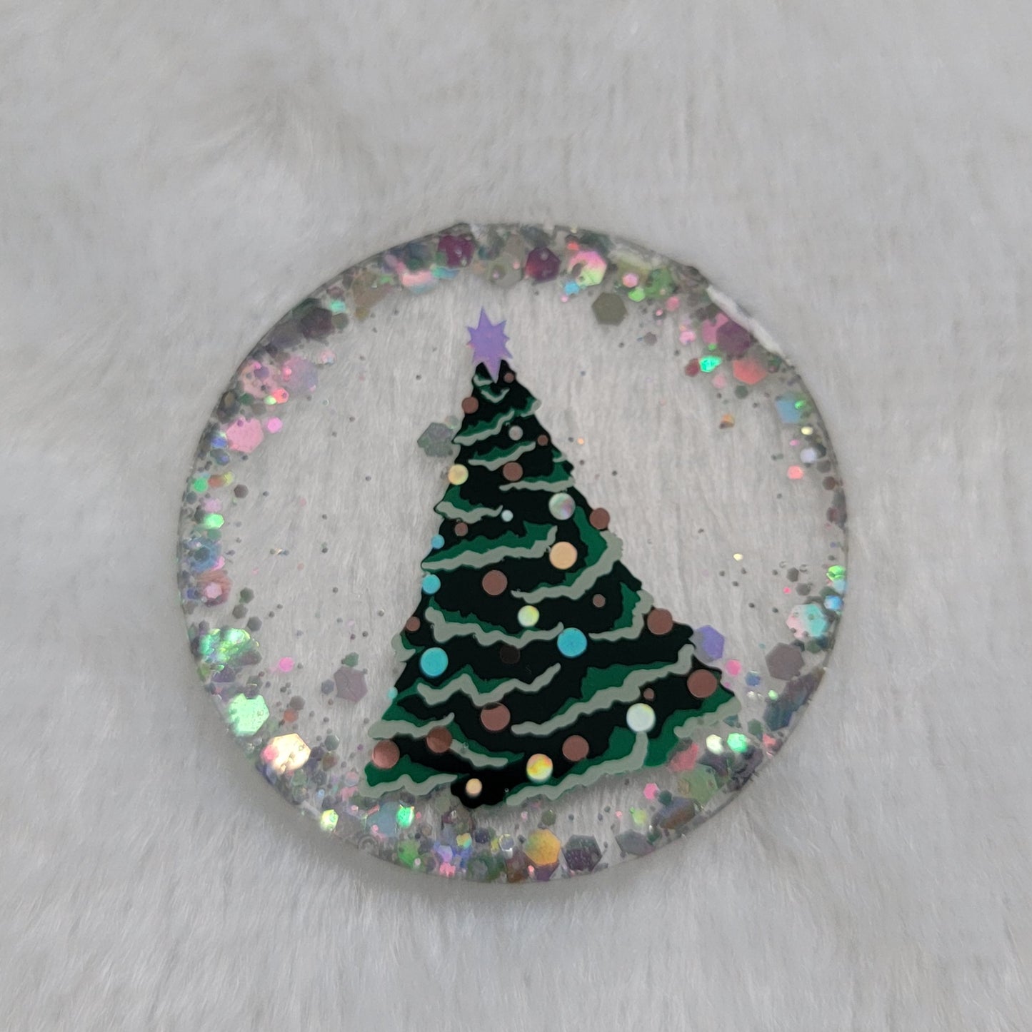 Round epoxy resin piece, base is transparent with silver holographic glitter around the edges, in the center there is a Christmas tree design made with vinyl in shades of green, silver and copper.