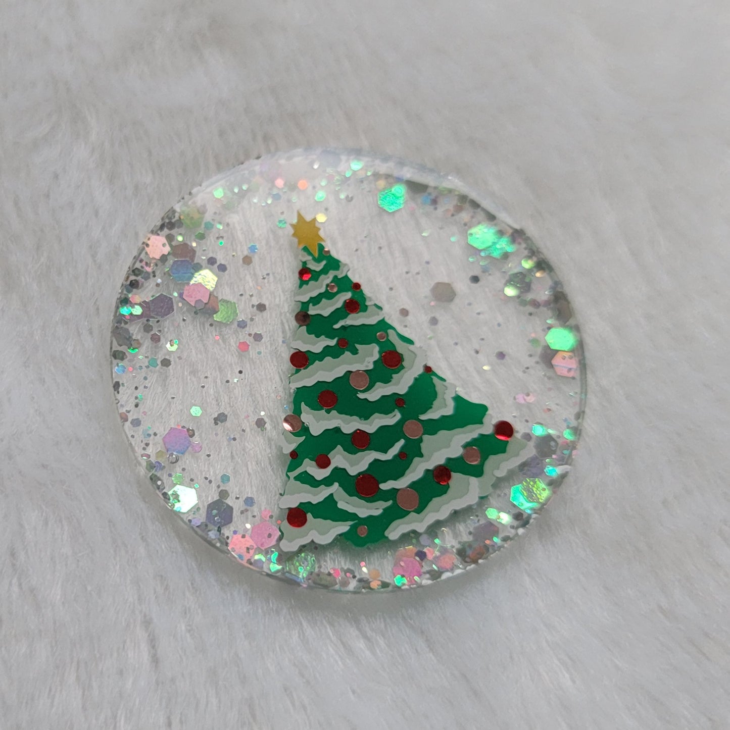 Round epoxy resin piece, base is transparent with silver holographic glitter around the edges, in the center there is a Christmas tree design made with vinyl in shades of green, white, red, copper and gold.