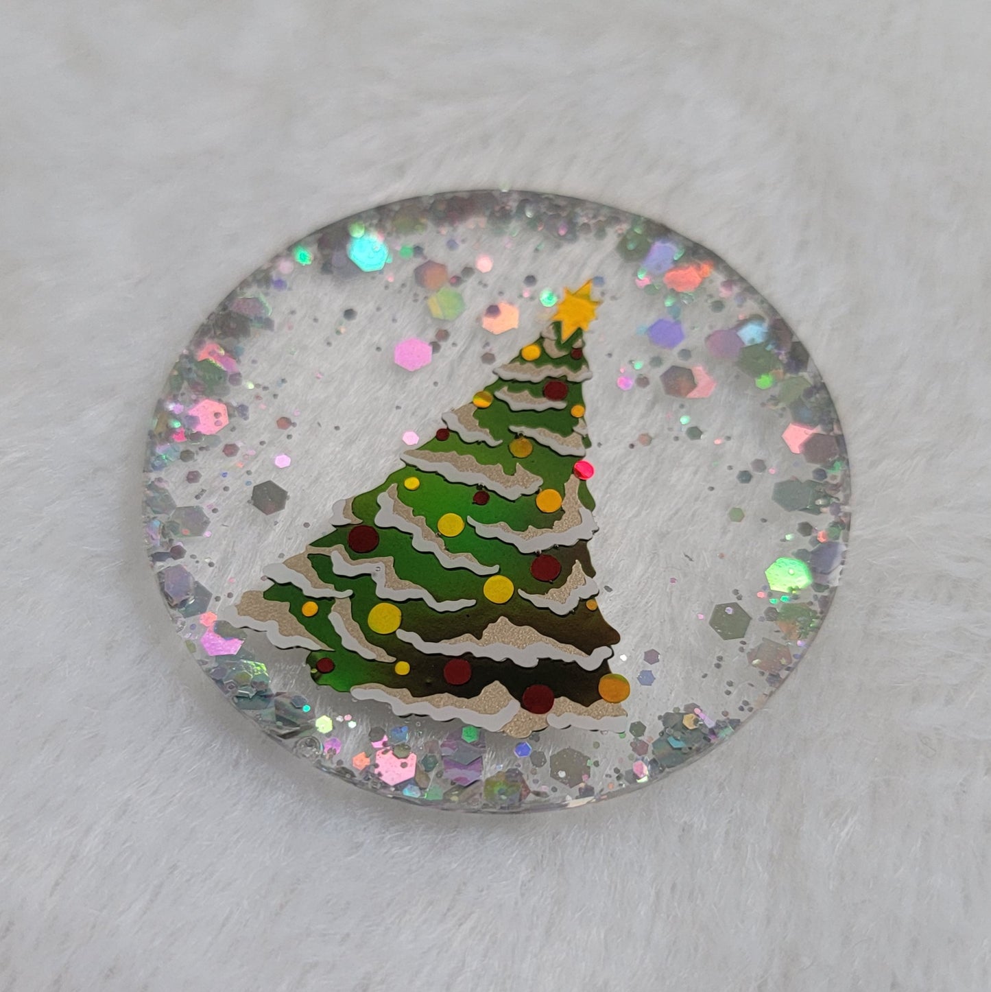 Round epoxy resin piece, base is transparent with silver holographic glitter around the edges, in the center there is a Christmas tree design made with vinyl in shades of green, white, red and gold.
