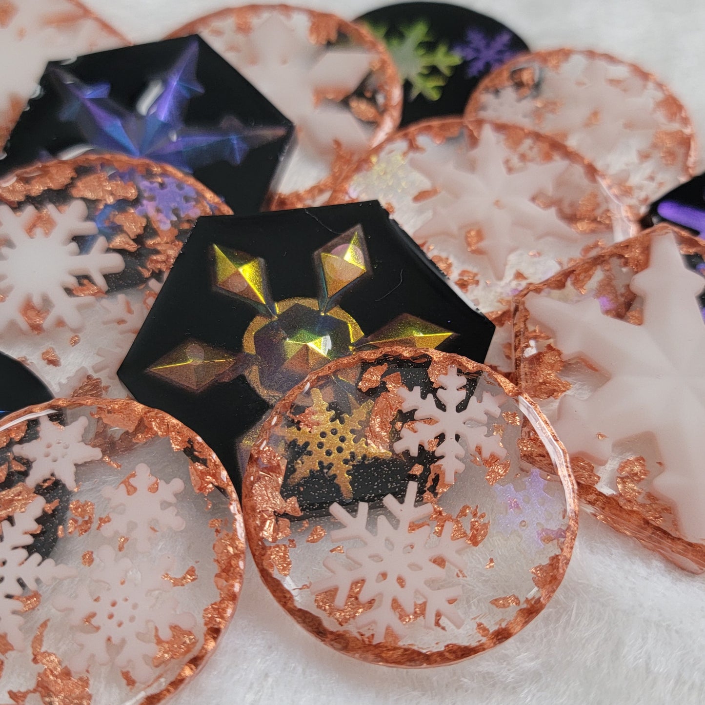 An array of epoxy resin pieces, hexagon shaped and round, containing small resin snowflake. Some with black backgrounds with iridescent snowflakes and some transparent with metallic copper accents and white snowflakes.