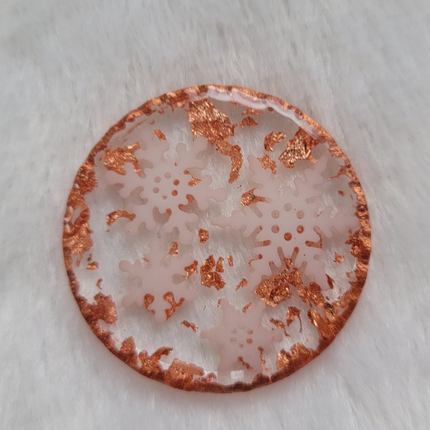 Round epoxy resin piece that contains four small white resin snowflakes, copper foil flakes, and has metallic copper edges that imitate hammered metal.