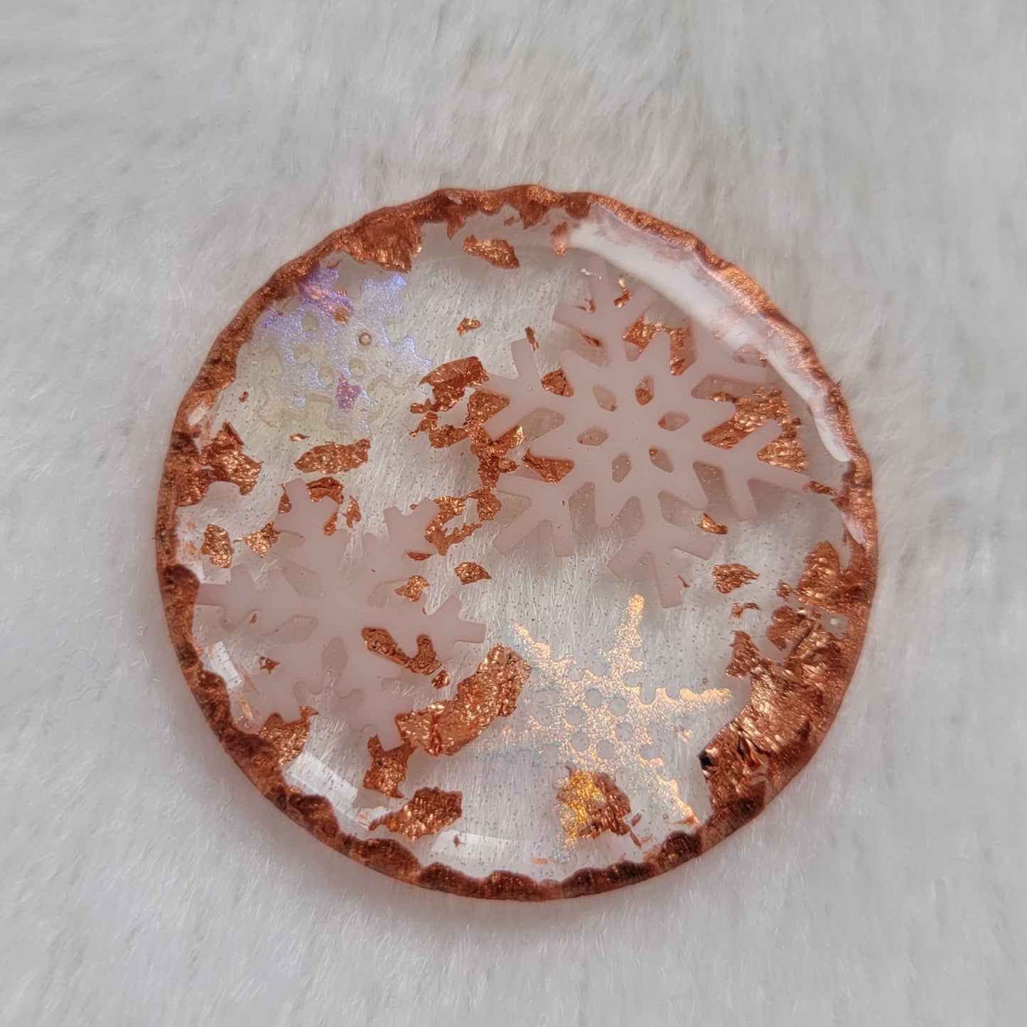 Round epoxy resin piece that contains four small resin snowflakes, two white and two translucent iridescent, copper foil flakes, and has metallic copper edges that imitate hammered metal.
