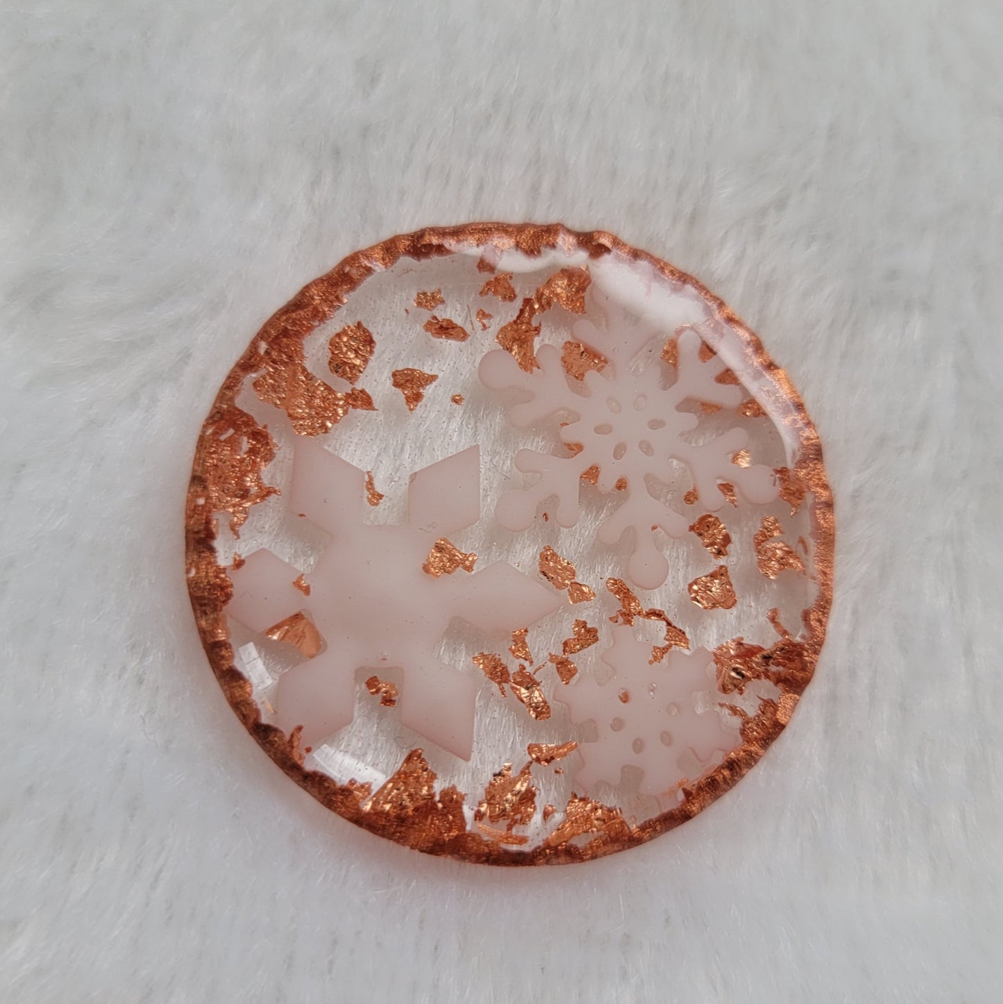 Round epoxy resin piece that contains three small white resin snowflakes, copper foil flakes, and has metallic copper edges that imitate hammered metal.