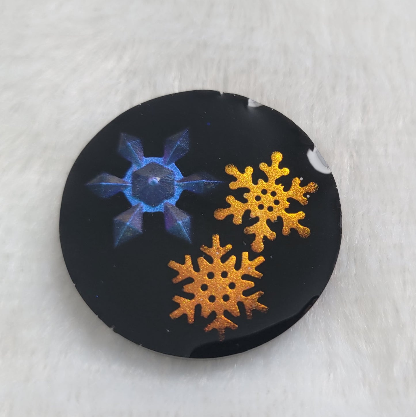 Round epoxy resin piece that contains three small resin snowflakes, one iridescent blue and two iridescent golden with a black background.