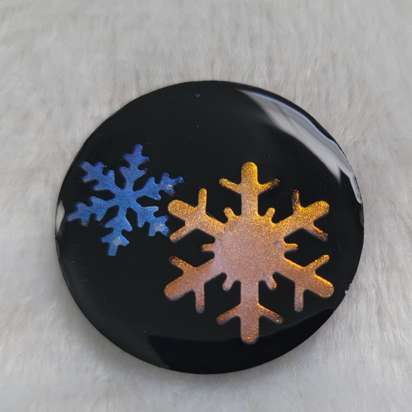 Round epoxy resin piece that contains two small resin snowflakes, one iridescent blue and one iridescent golden with a black background.