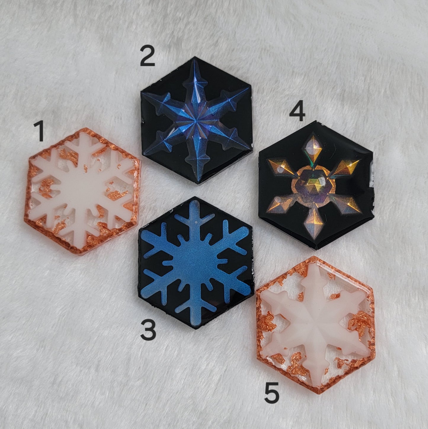 Five hexagon shaped epoxy resin pieces, three with iridescent resin snowflakes and black backgrounds and two with white snowflakes and copper accents.