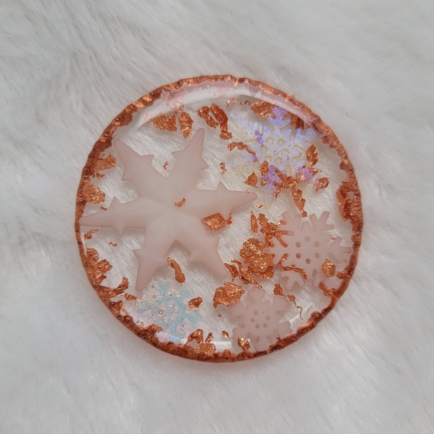 Round epoxy resin piece that contains five small white resin snowflakes, three white and two translucent iridescent, copper foil flakes, and has metallic copper edges that imitate hammered metal.