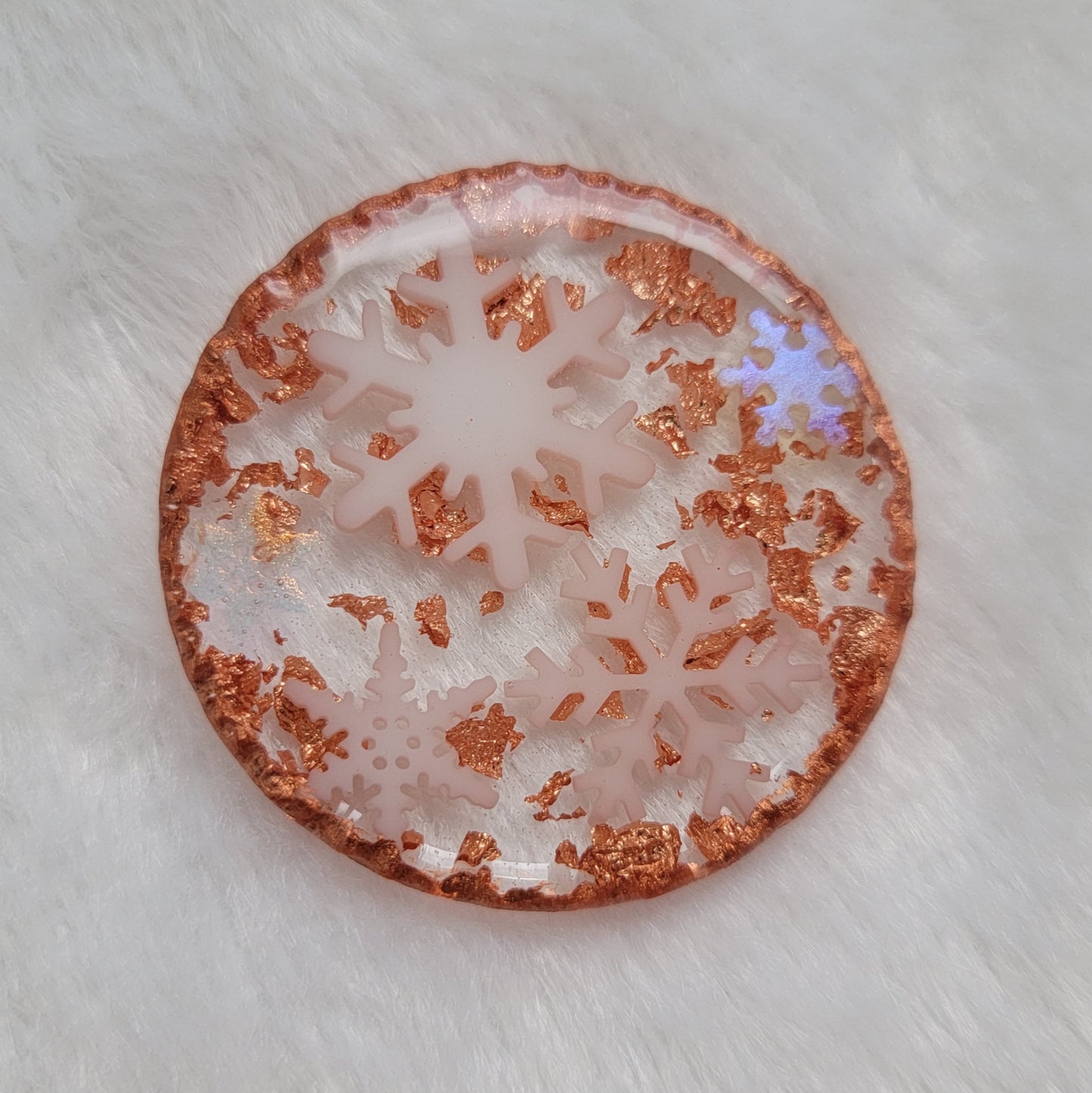 Round epoxy resin piece that contains five small white resin snowflakes, three white and two translucent iridescent, copper foil flakes, and has metallic copper edges that imitate hammered metal.