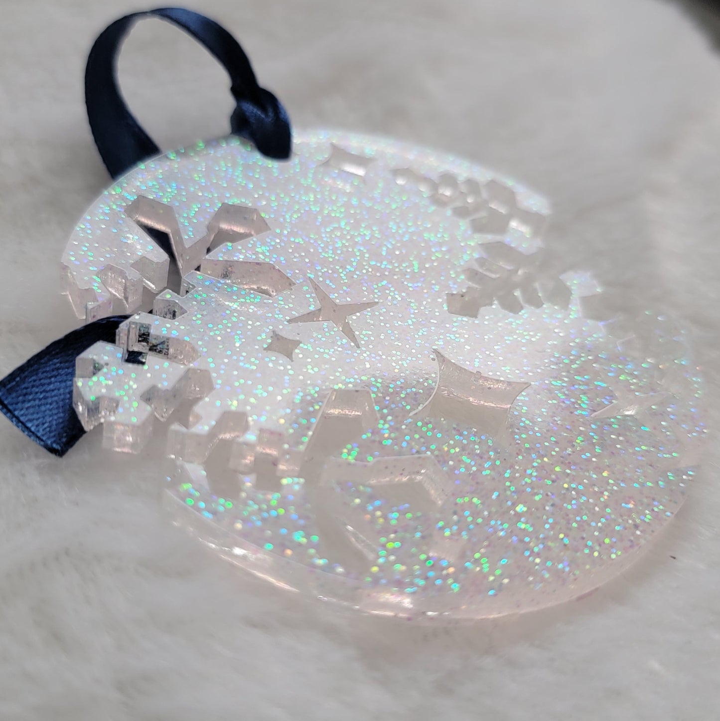 A round epoxy resin ornament that has snowflake shapes cutout from each side, made with iridescent white glitter and a dark blue ribbon attached.