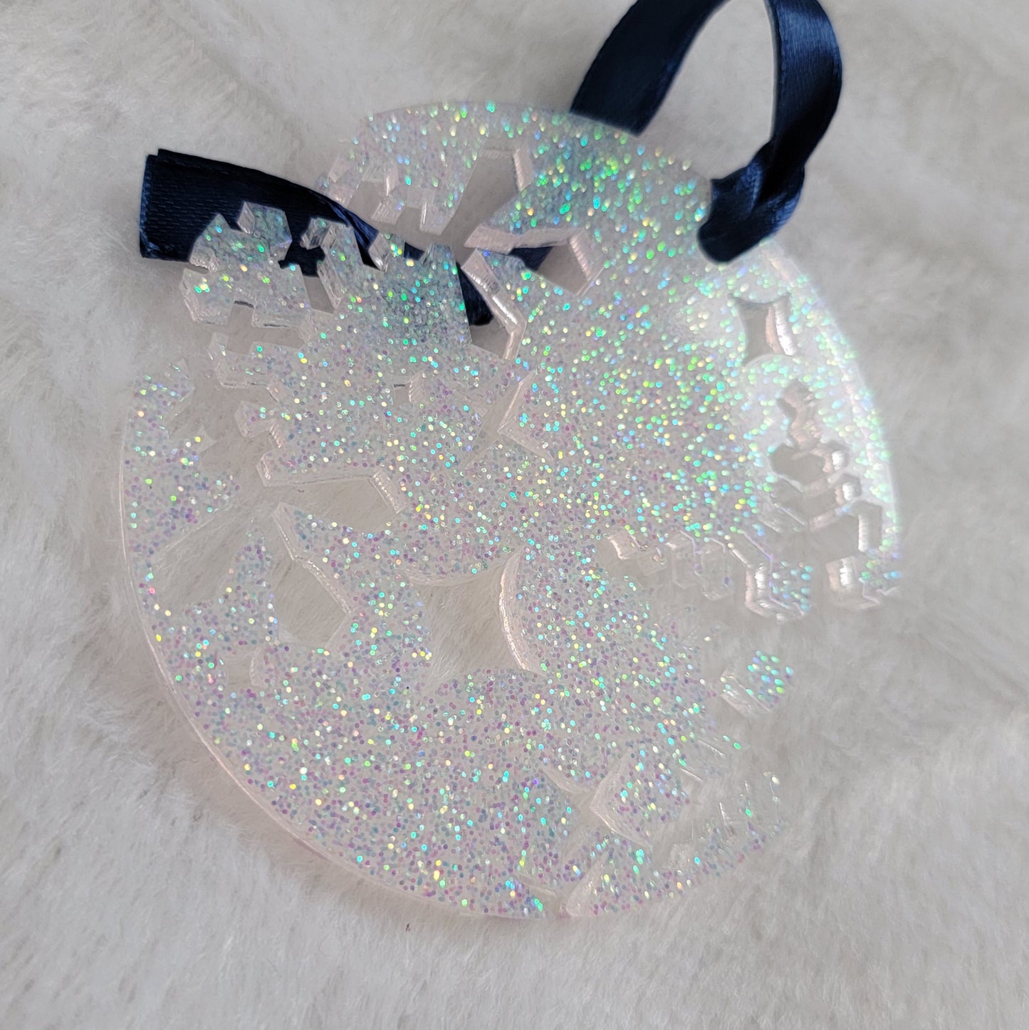 A round epoxy resin ornament that has snowflake shapes cutout from each side, made with iridescent white glitter and a dark blue ribbon attached.