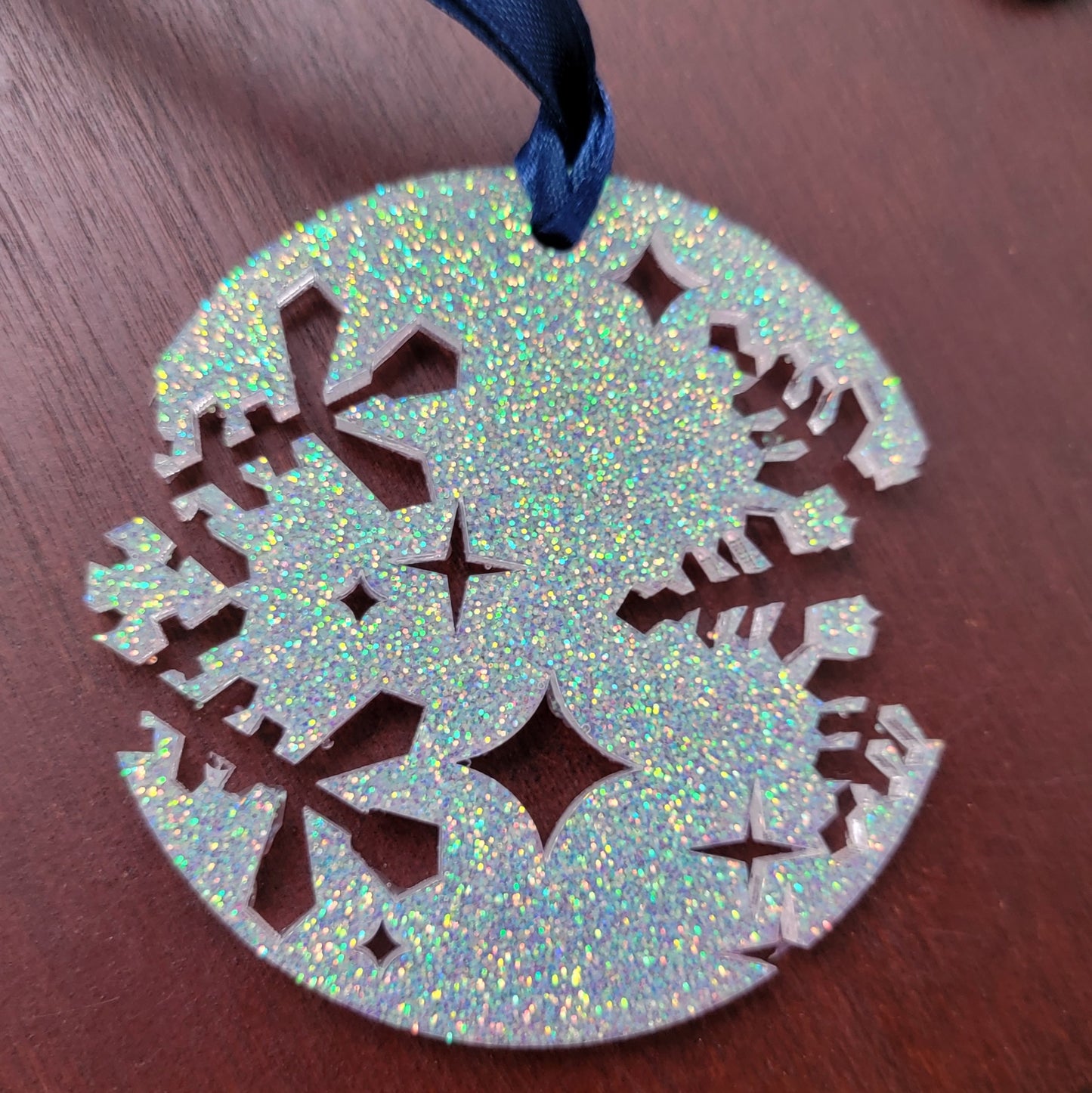 A round epoxy resin ornament that has snowflake shapes cutout from each side, made with iridescent white glitter and a dark blue ribbon attached.