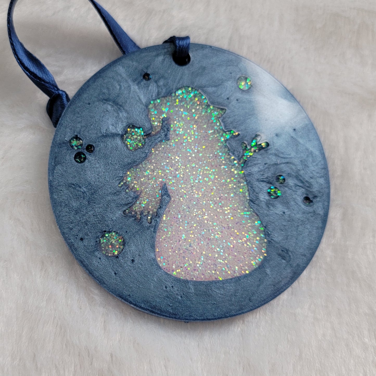 A round epoxy resin ornament that has an iridescent white glitter snowman silhouette in the middle, made with ice blue pigment and a dark blue ribbon attached.