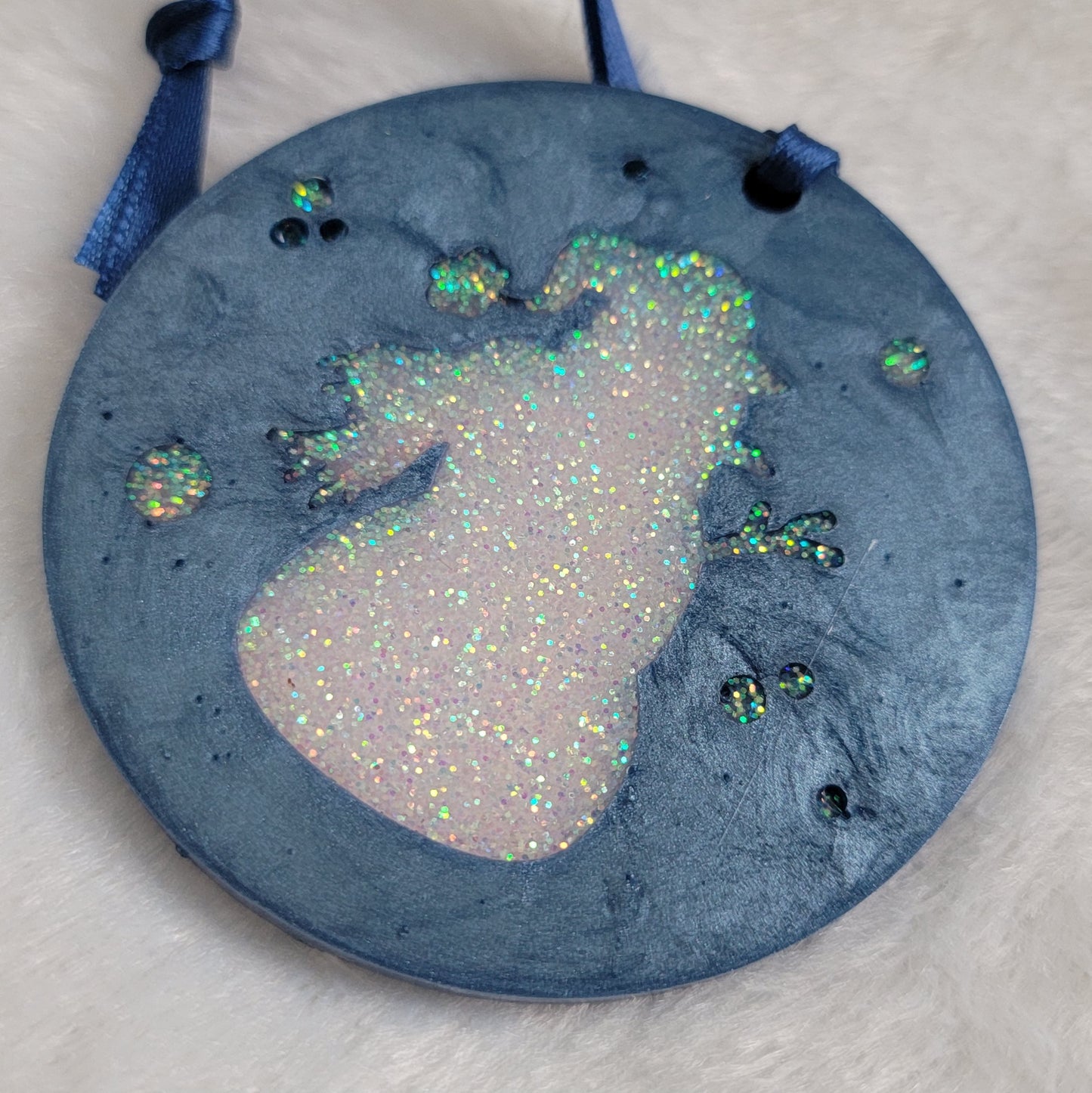A round epoxy resin ornament that has an iridescent white glitter snowman silhouette in the middle, made with ice blue pigment and a dark blue ribbon attached.