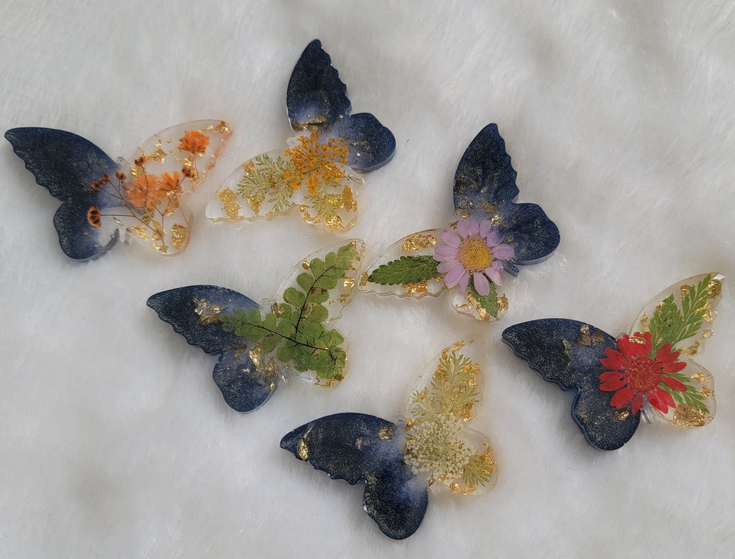 Six butterfly shaped epoxy resin pieces, made with dark shining blue pigment, golden foil flakes and varies dried flowers and foliage.