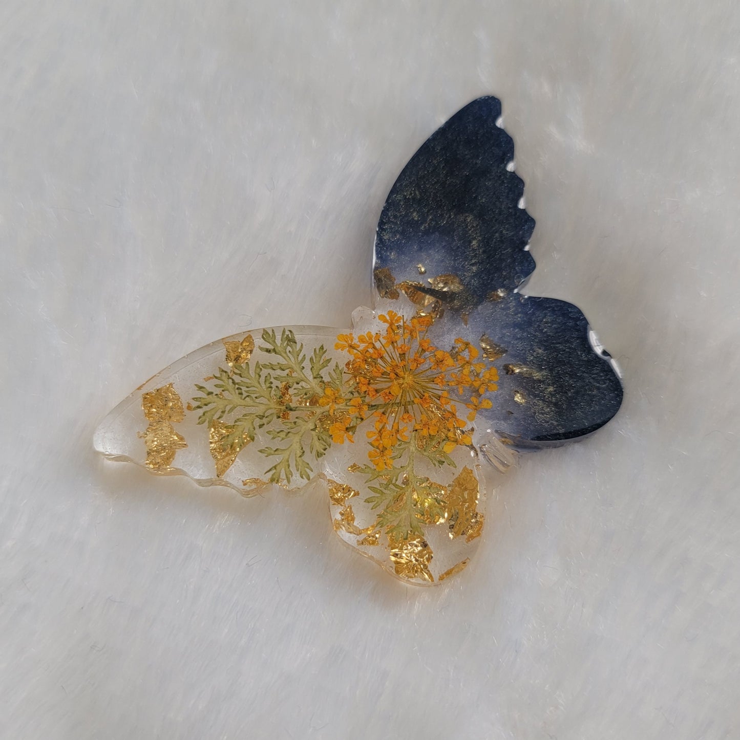 Butterfly shaped epoxy resin piece, right wings dark shimmering blue pigment and left wings contain golden foil flakes, foliage and small orange flower.