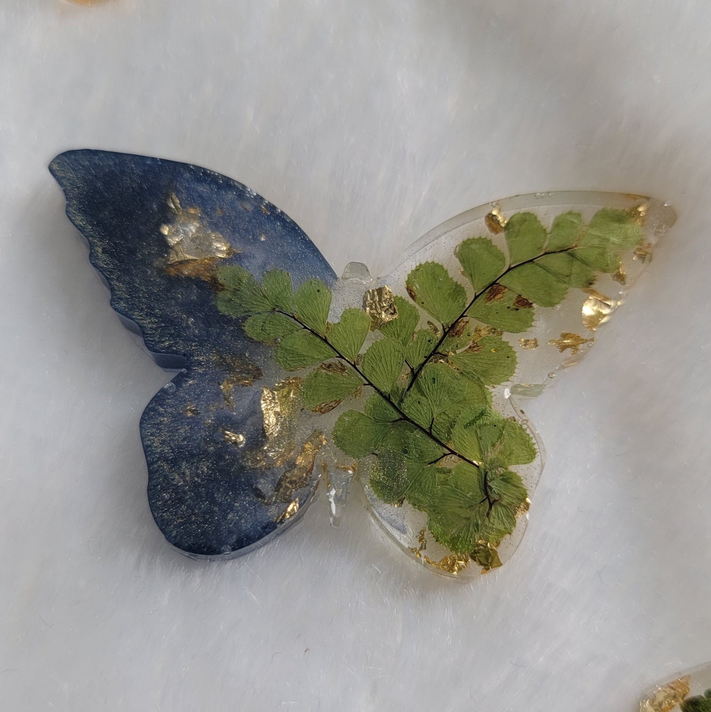 Butterfly shaped epoxy resin piece, left wings dark shimmering blue pigment and right wings contain golden foil flakes and green foliage.