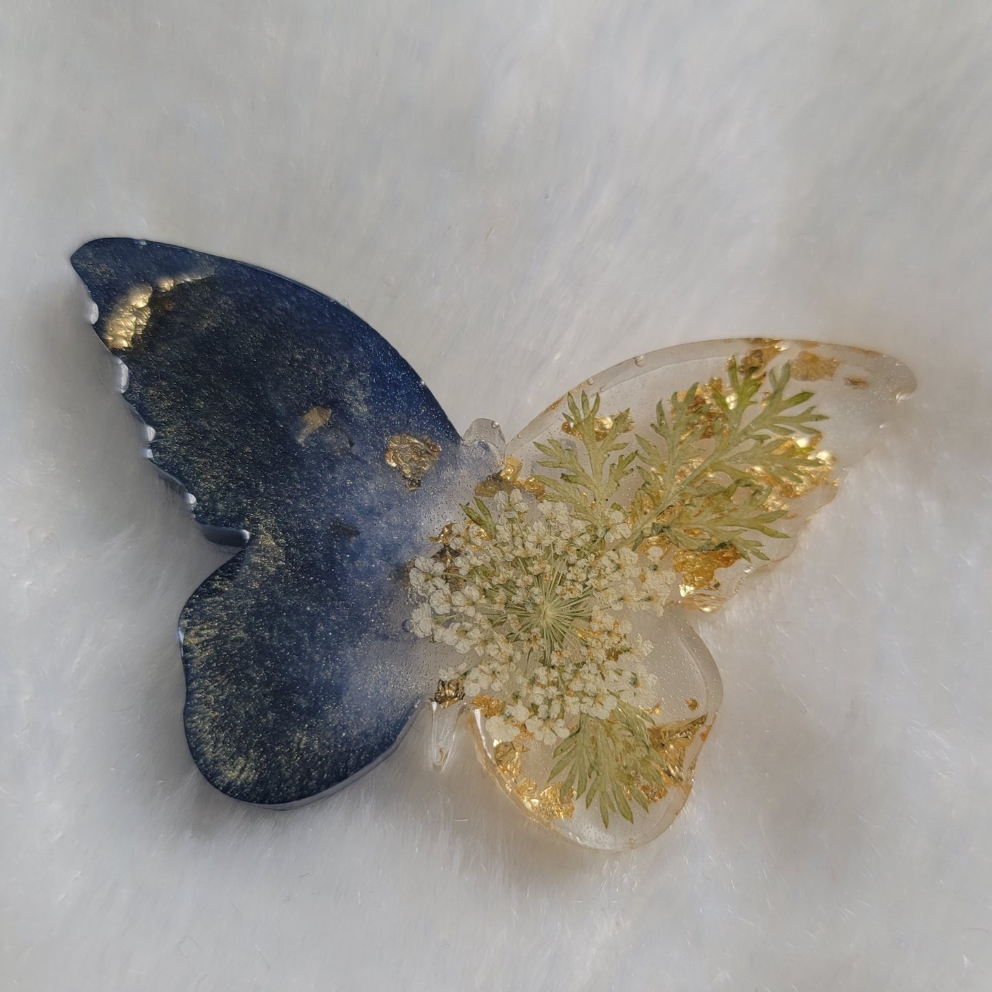Butterfly shaped epoxy resin piece, left wings dark shimmering blue pigment and right wings contain golden foil flakes, foliage and small white flower.