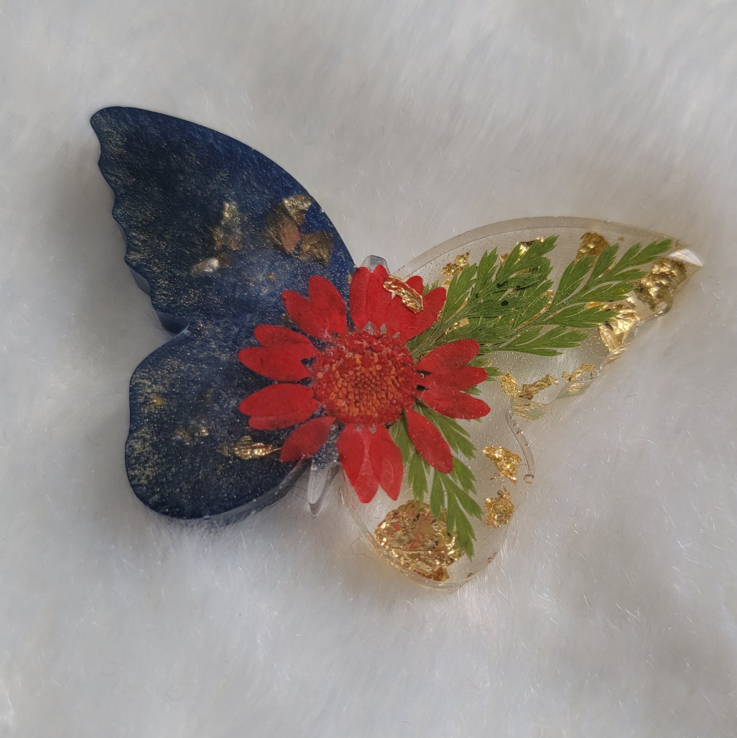 Butterfly shaped epoxy resin piece, left wings dark shimmering blue pigment and right wings contain golden foil flakes, foliage and red daisy flower.