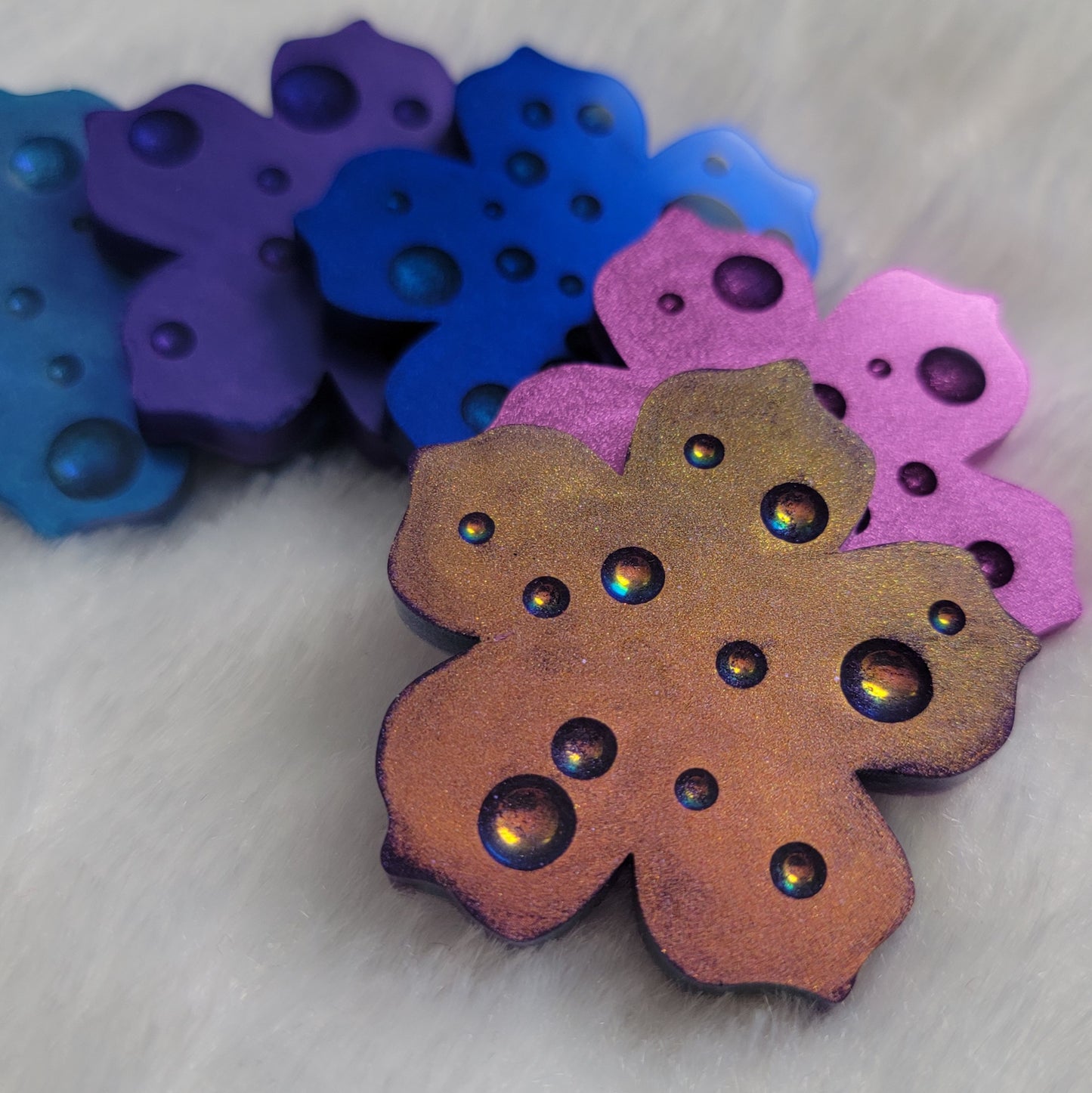 Five flower blossom shaped epoxy resin pieces with dewdrop effect and shimmering metallic finishes in shades of purple and blue.