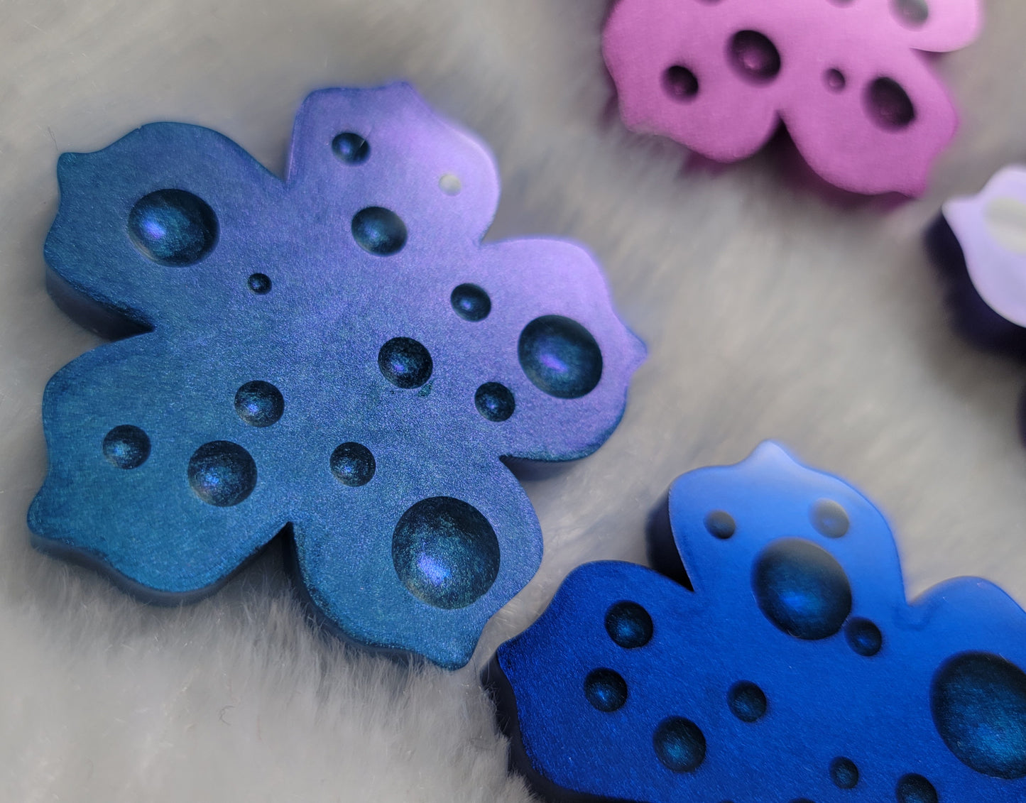 Flower blossom shaped epoxy resin pieces with dewdrop effect and shimmering metallic finishes in shades of purple and blue.