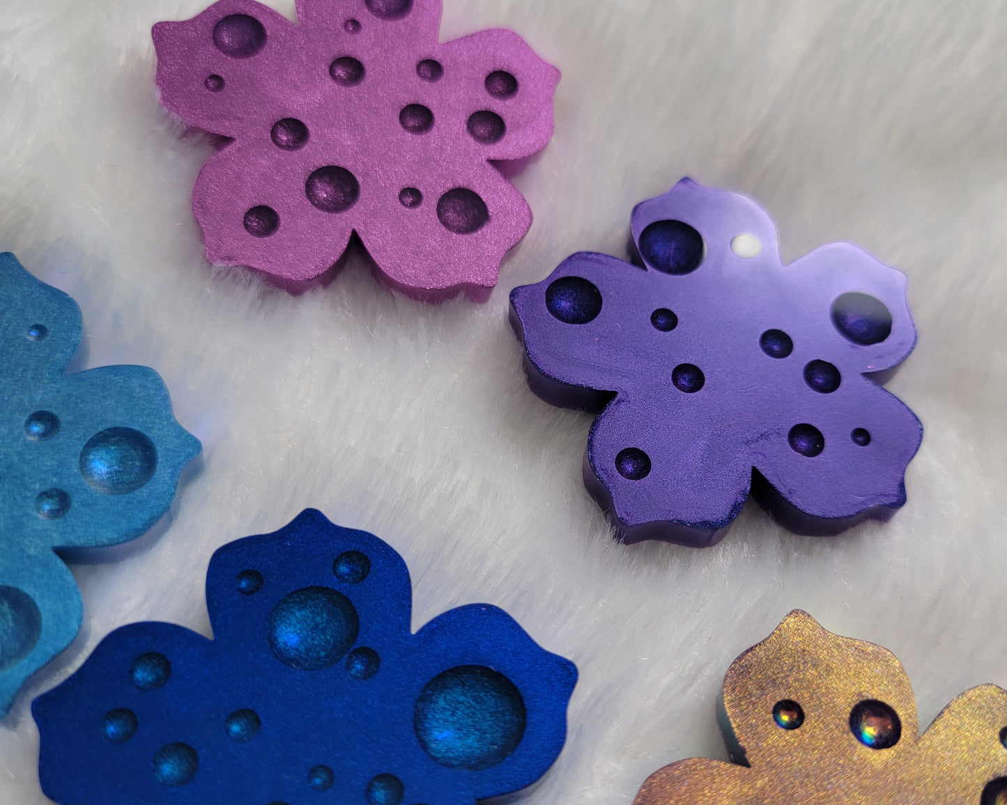 Flower blossom shaped epoxy resin pieces with dewdrop effect and shimmering metallic finishes in shades of purple and blue.