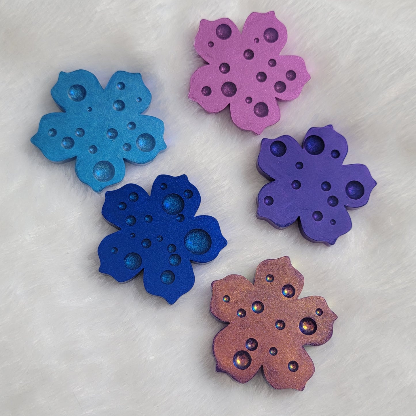 Five flower blossom shaped epoxy resin pieces with dewdrop effect and shimmering metallic finishes in shades of purple and blue.